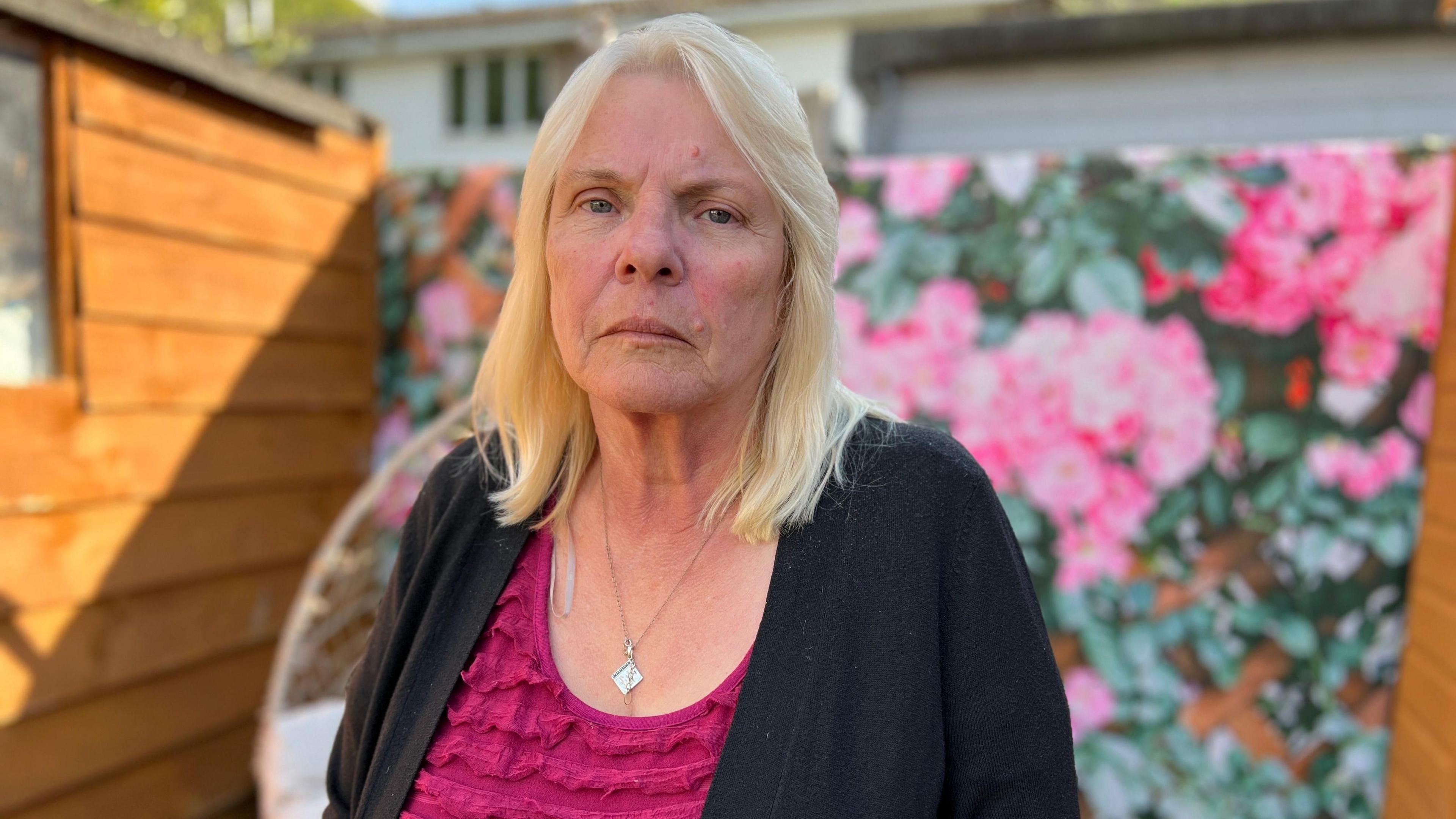 Mary Richardson, who is wearing a pink top and black cardigan. She is in a garden and looking seriously at the camera.