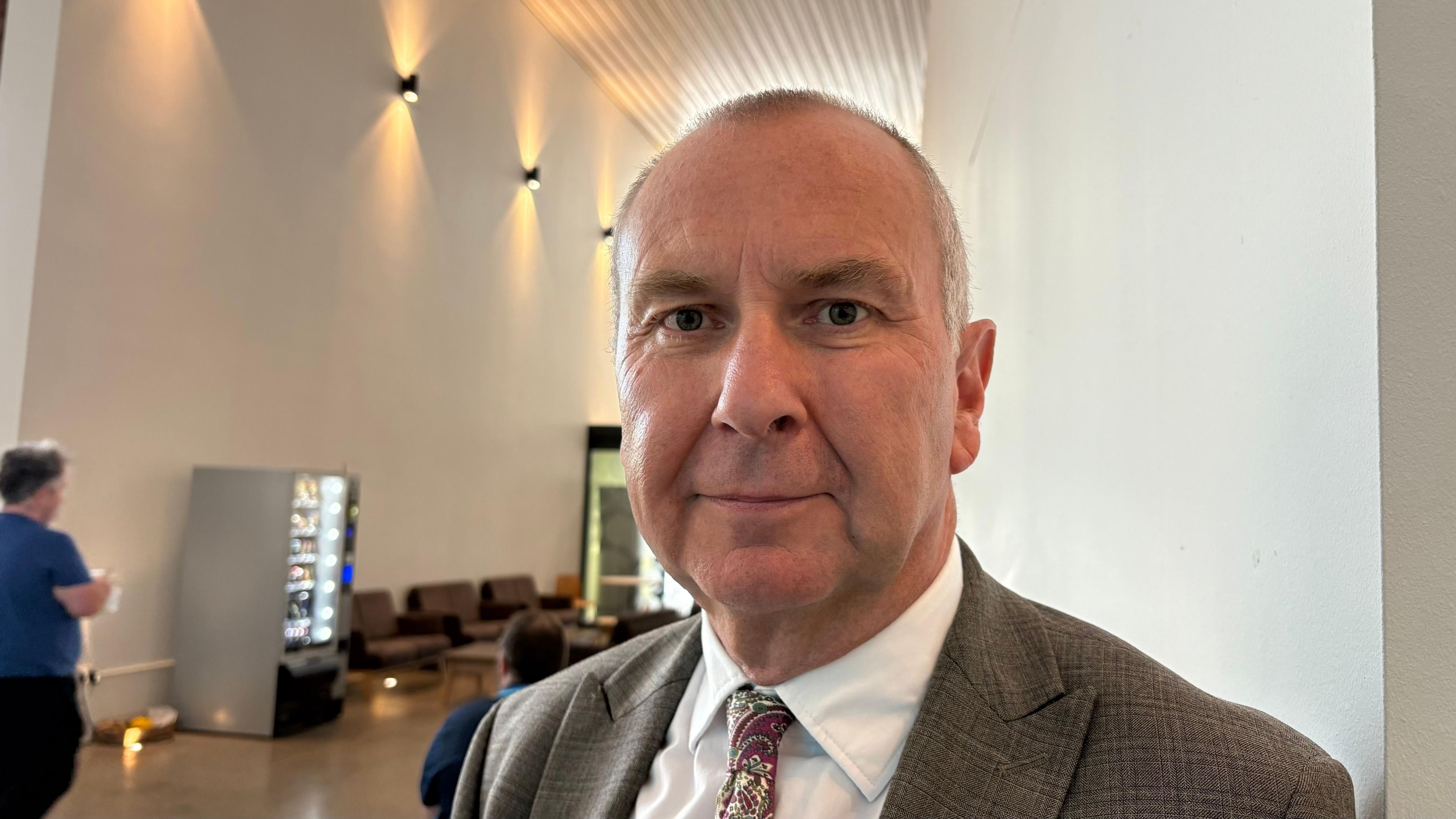 Steve Kelly with short white hair, brown jacket and red tie looking at the camera with a cafe in the background