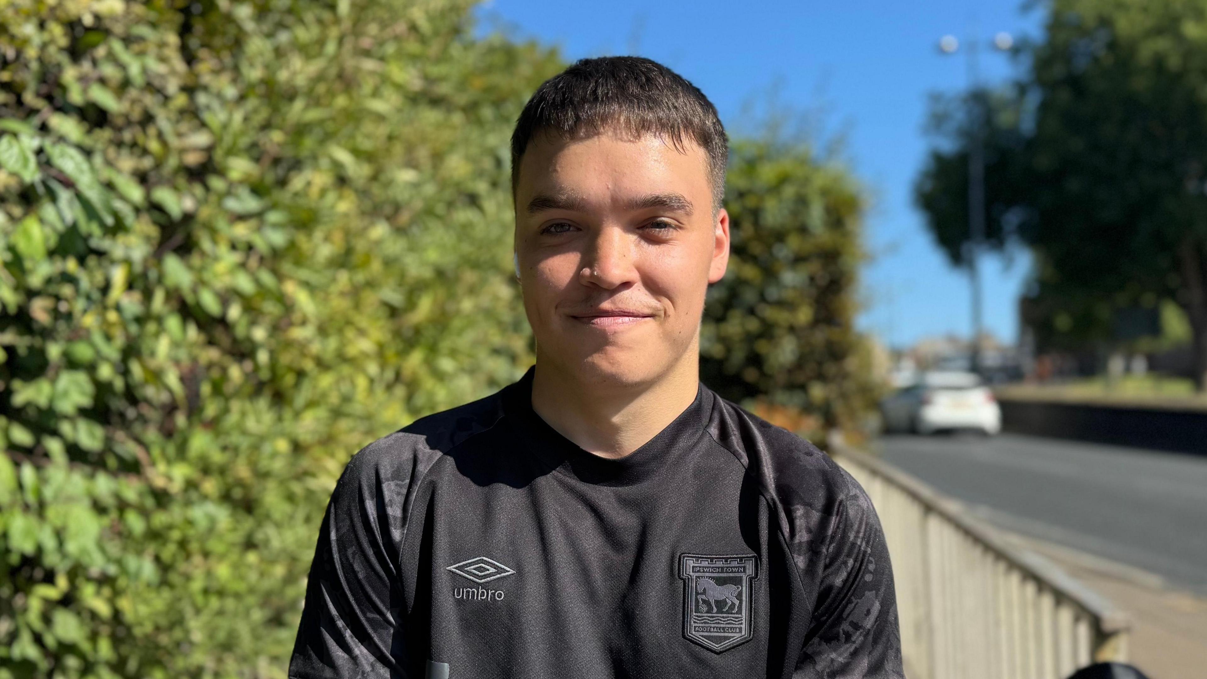 A head and shoulders image of Harry Ward. He is wearing a black Ipswich Town top and smiling into the camera. A road and a tree can be seen in the background.