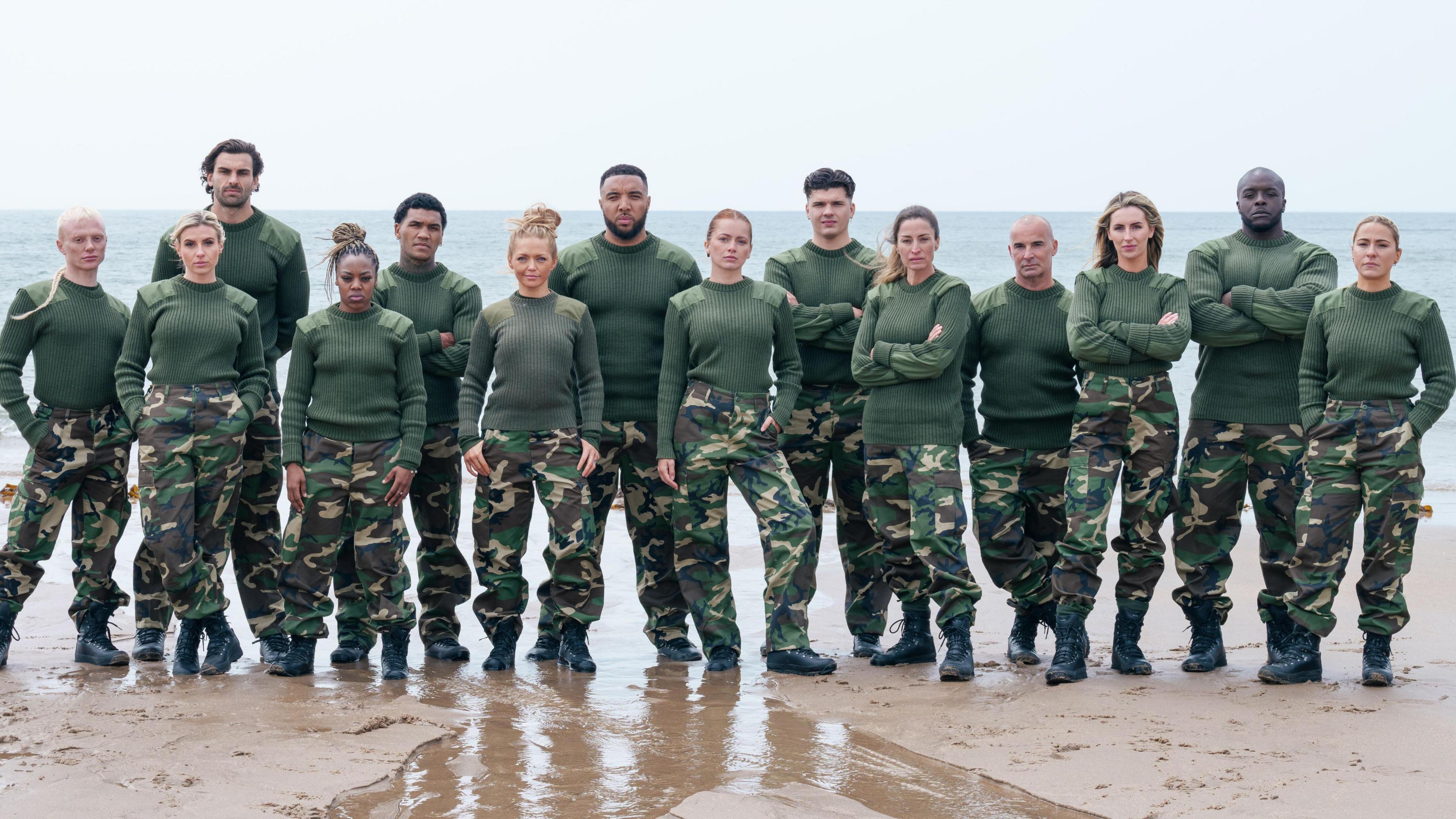 The celebrity recruits, wearing military fatigues, line up on a beach with their backs to the sea. They are stood in two rows, some with their arms crossed, with serious expressions.
