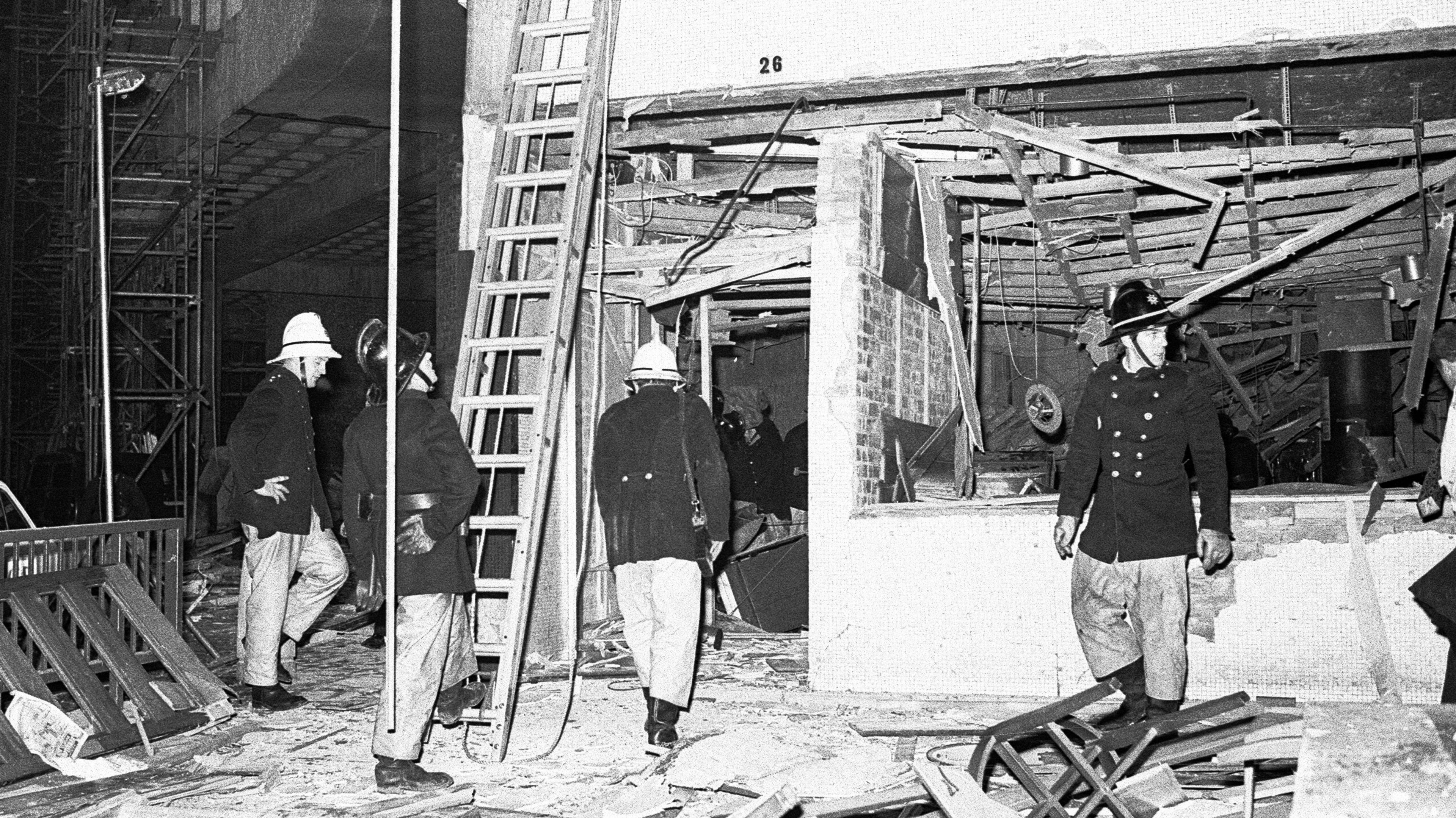 Black and white image of four policemen inspecting the wreckage of a bombed-out building. They are surrounded by broken bits of furniture and rubble. They are all wearing old-fashioned policeman's helmets, jackets with buttons down the front, trousers and boots.