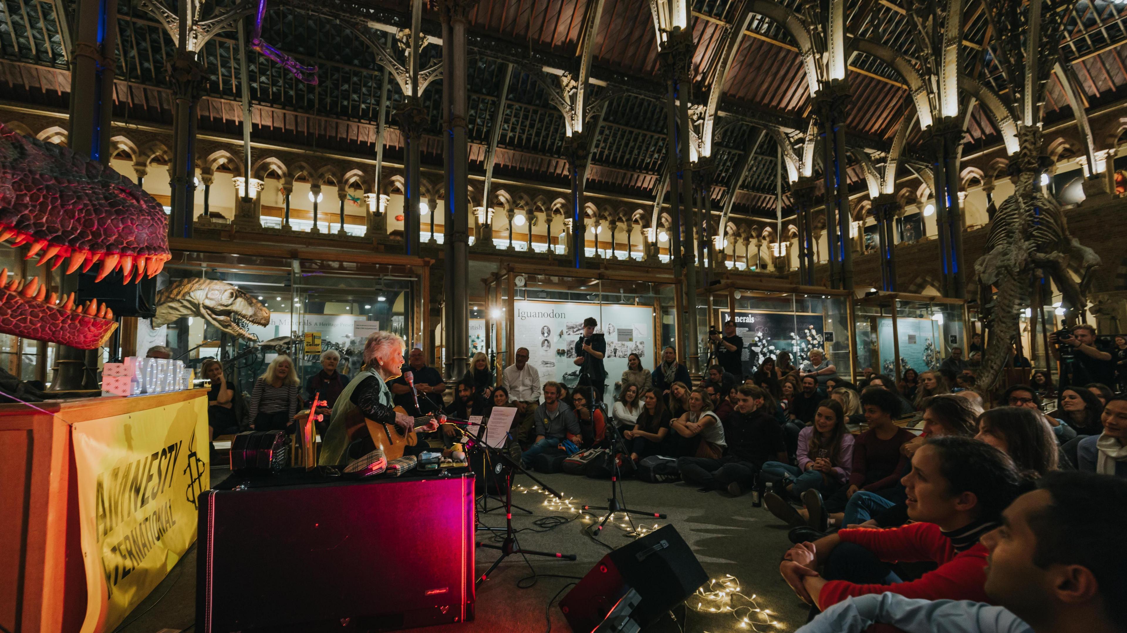 Peggy Seeger at the Museum of Natural History in Oxford where the audience is sitting on the ground in front of her. Museum artefacts can be seen around them such as dinosaur reconstructions.