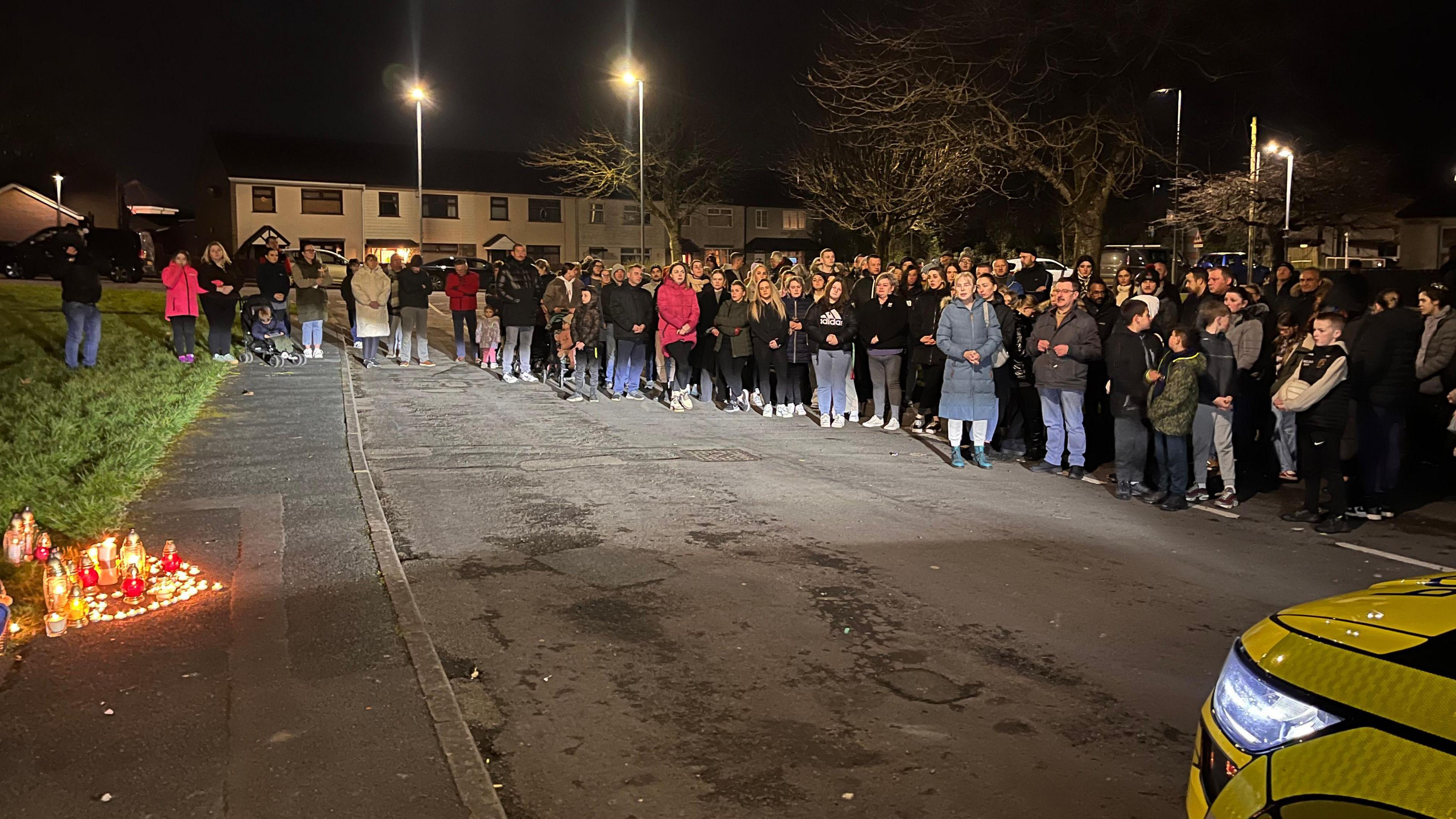 Mourners attending a vigil for the victim in the Lisnahull Estate on Friday night.  A large crowd of people are standing close together for the ceremony, many with their hands clasped in prayer.   A number of lit candles are placed on a pavement in front of them.  The front of a parked police vehicle is partially visible in the foreground.