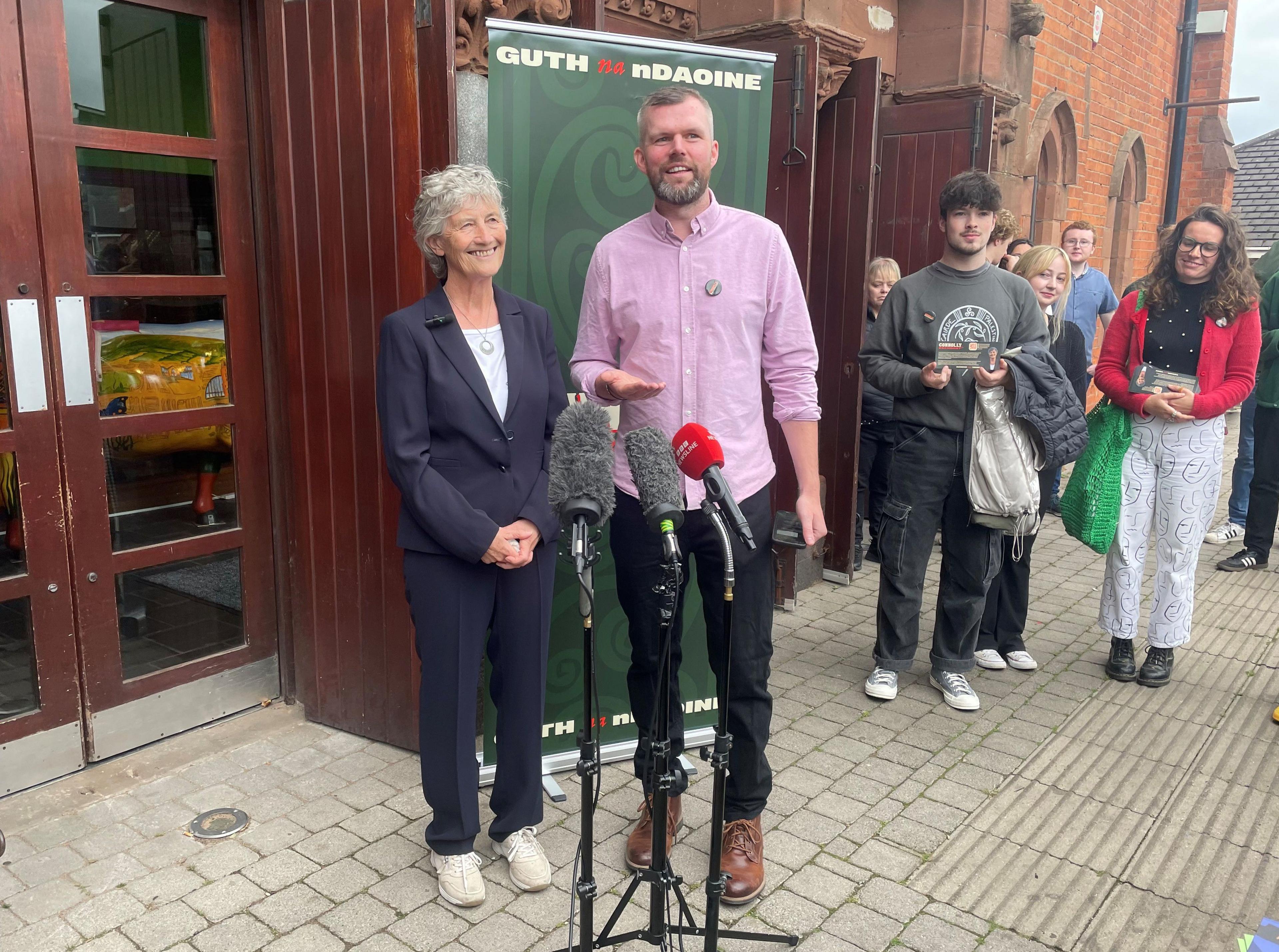 Connolly, pictured in a navy trouser suit, she has short grey hair and is stood alongside People Before Profit MLA Gerry Carroll, who is in black trousers brown boots and a pink shirt, during a visit to Culturlann McAdam O Fiach in Belfast as part of the event facilitated by People Before Profit.
They are stood outside a brown wooden building, with young people pictured in the background