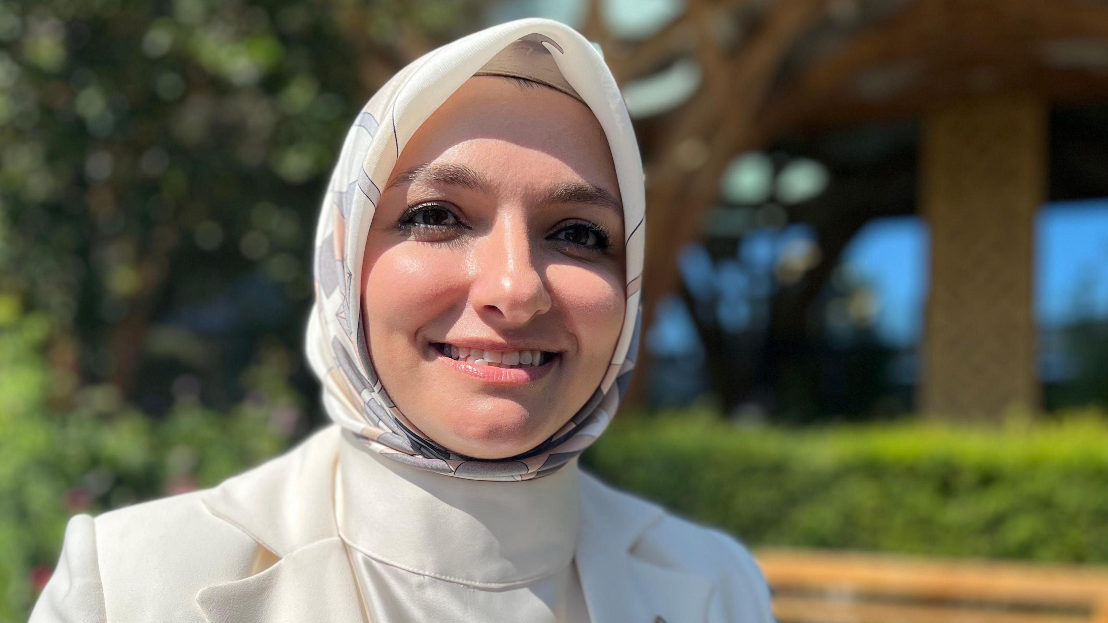 Zeynep Coskun is standing in front of trees which are outside a wooden building. She is wearing a white patterned hijab and is smiling. She is wearing a white T-shirt and jacket.