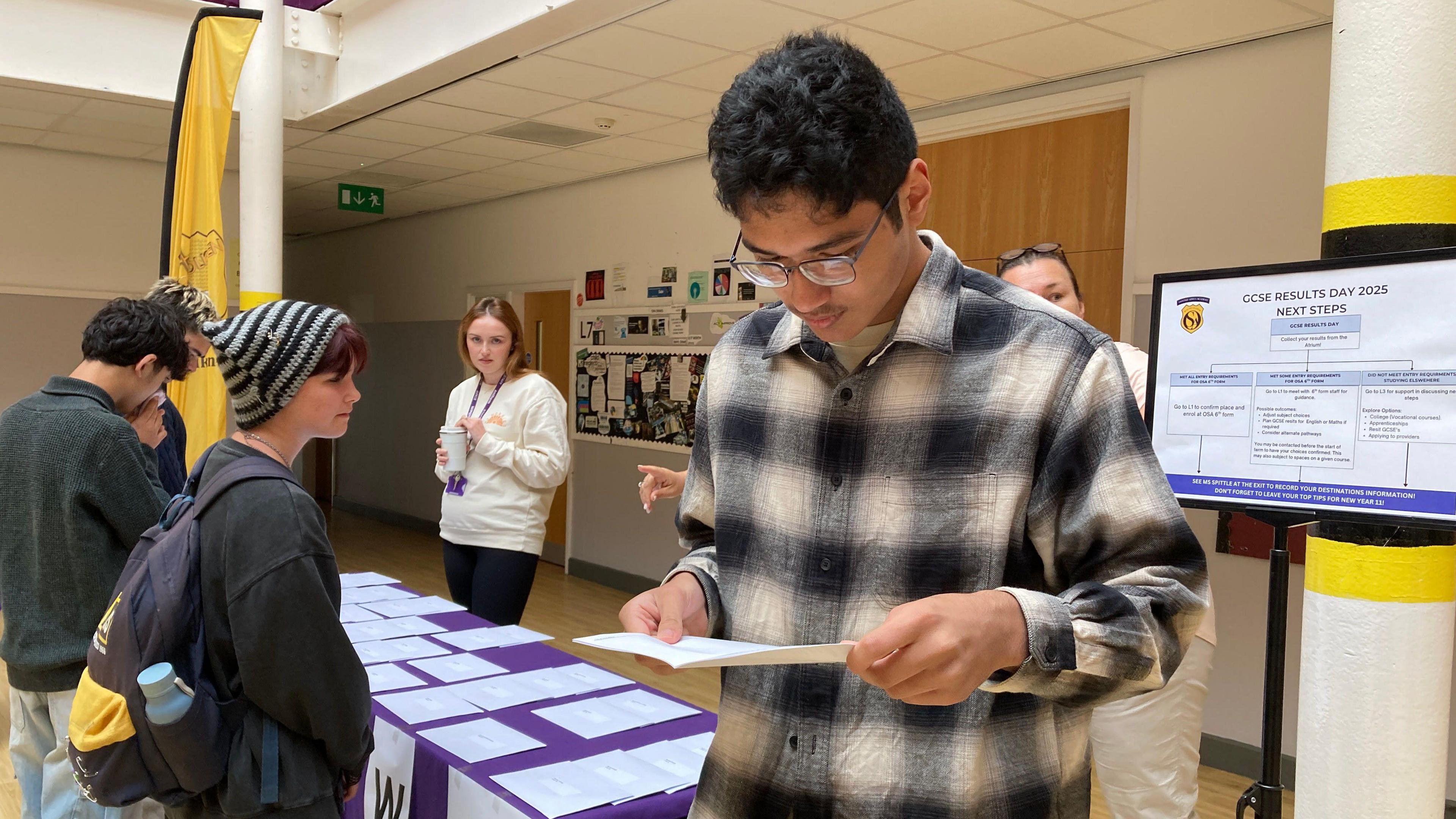 A student is standing looking down at his results. He has thin black wire framed glasses and short black hair. He is wearing a black and beige checked long sleeve shirt. In the background behind him other students are waiting at a table covered in white envelopes to collect their results.