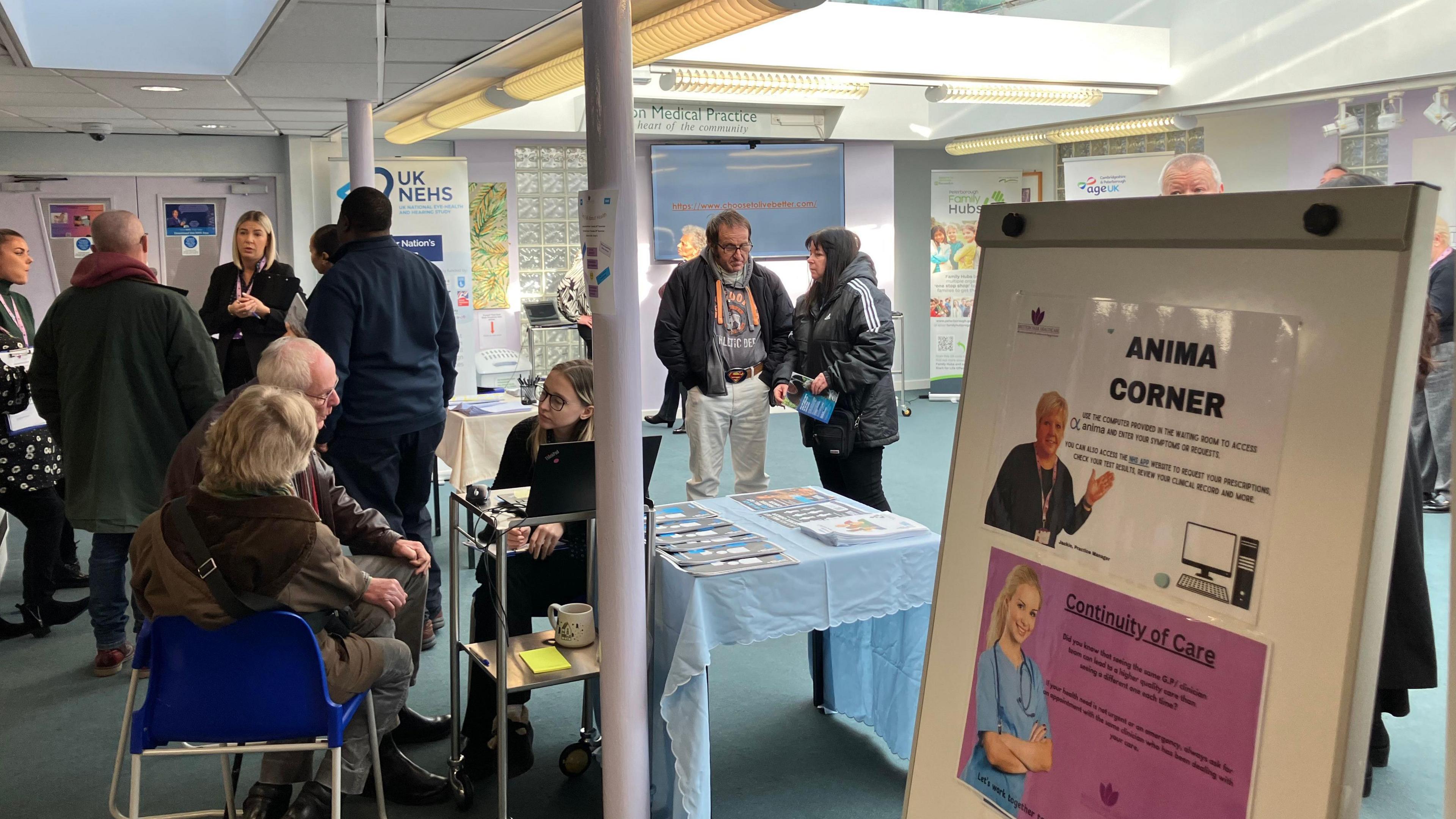 Members of public inside the surgery room with blue carpets - some sitting on blue chairs, others standing around, with posters of information placed across the room.