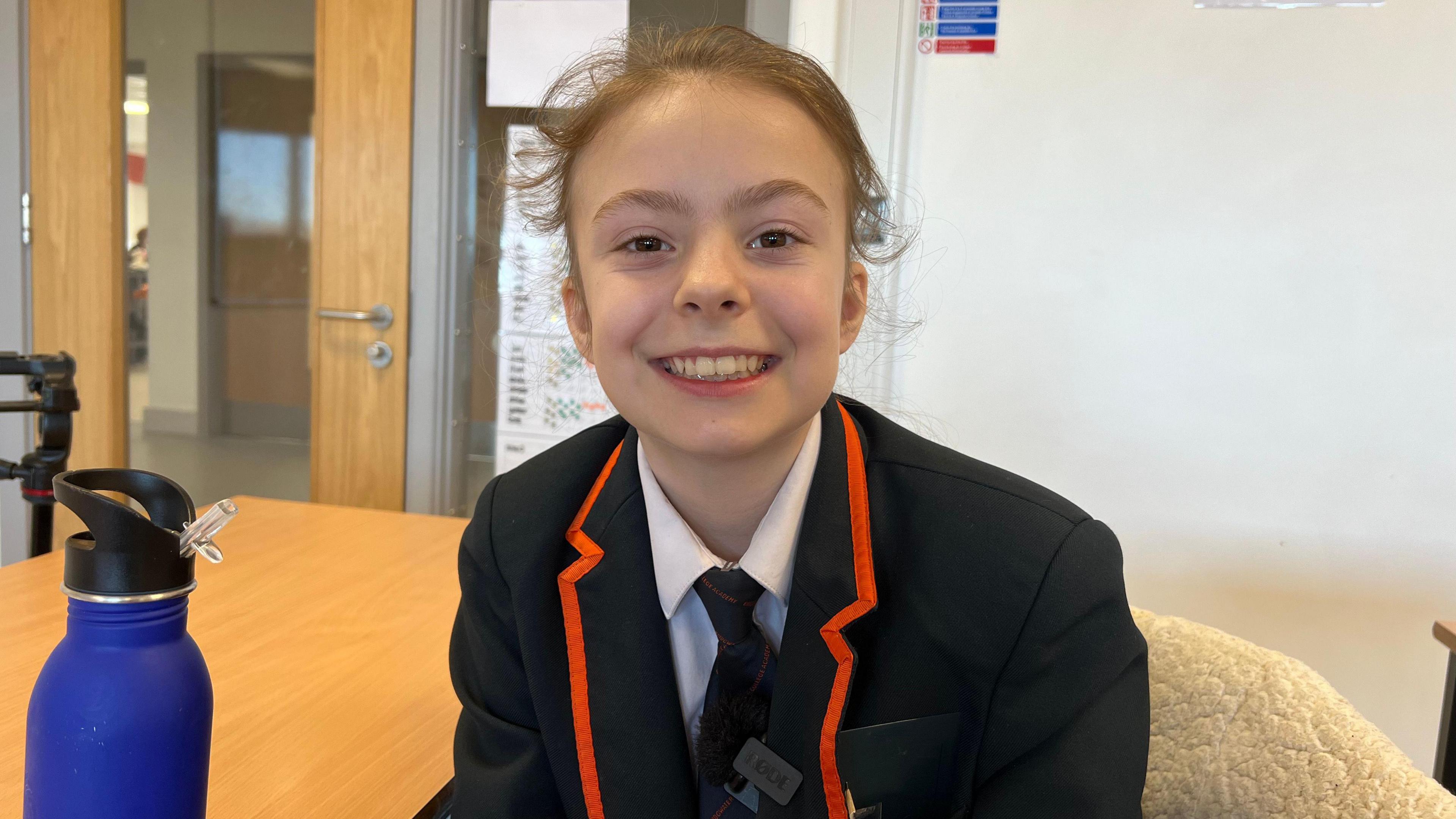 A female pupil smiles at the camera. She is wearing a dark blazer, white shirt and dark tie and is in an empty classroom