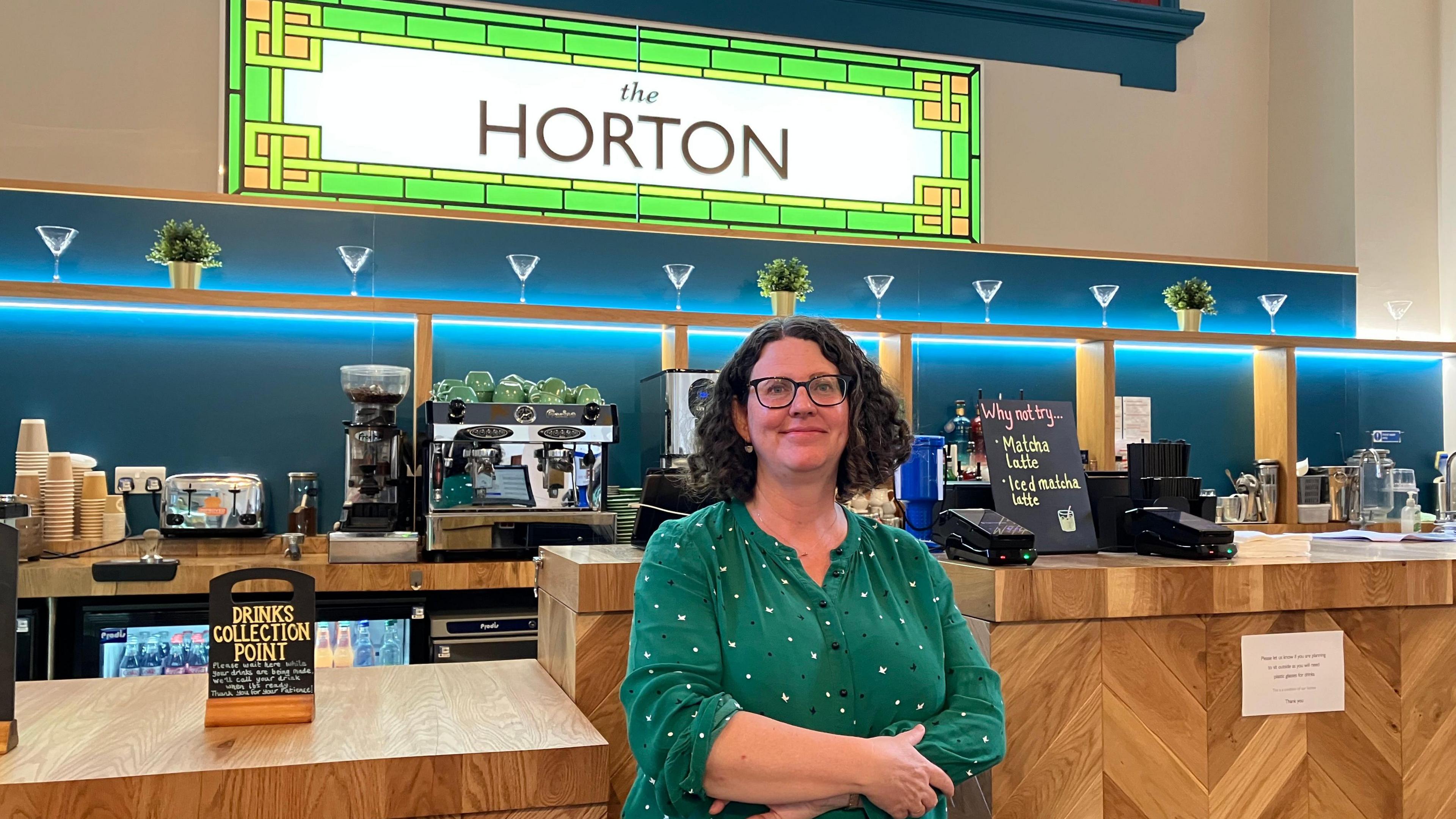 Prof Alana Harris stands in front of a wooden bar with a green stained glass sign behind her which is illuminated and reads "the Horton". She is looking at the camera and smiling, and has short brown curly hair.