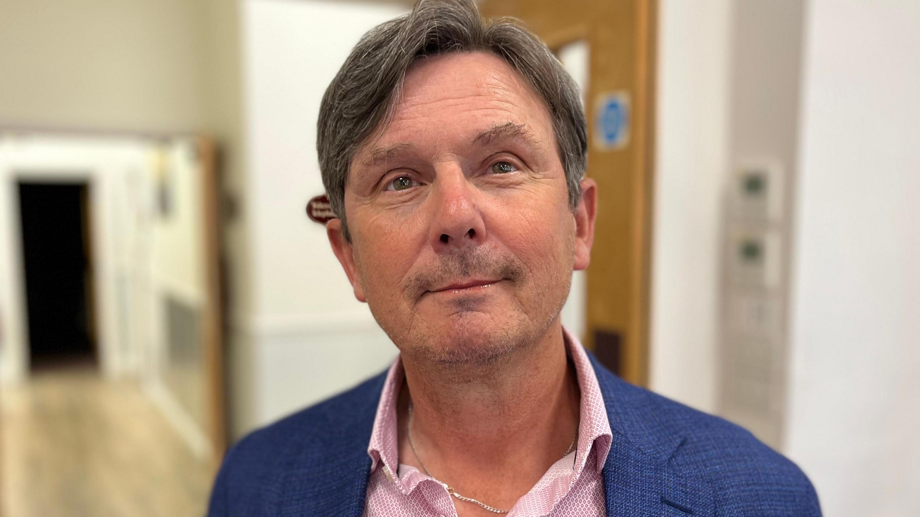 Deputy Andy Sloan - A man with black and grey hair.  He is wearing a blue suit, over a pink shirt. 