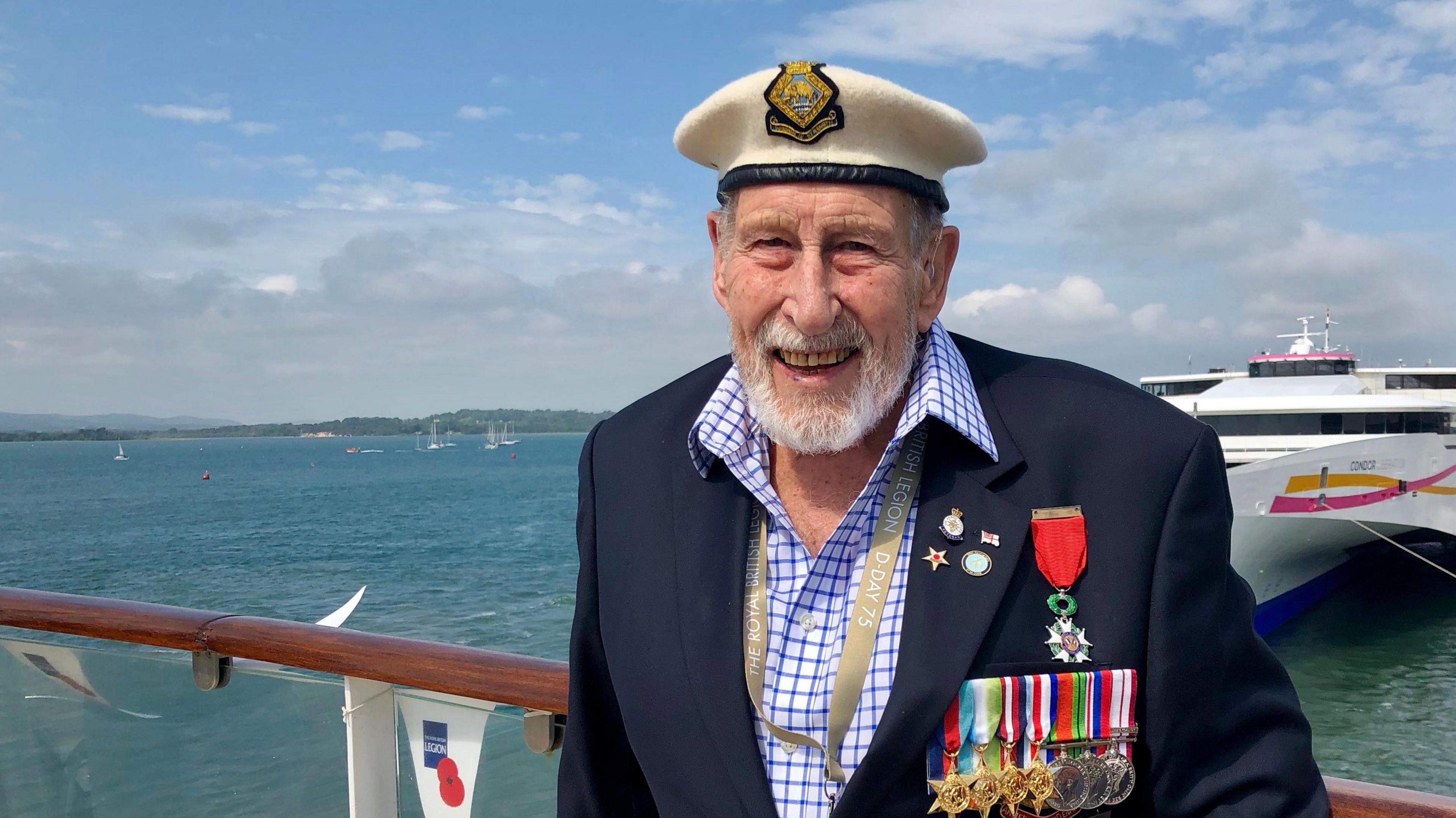 George Winter from shoulders up wearing a suit adorned in medals and wearing a sailors hat. He is stood on a boat, with other boats in the background as well as the water and shoreline in the distance.