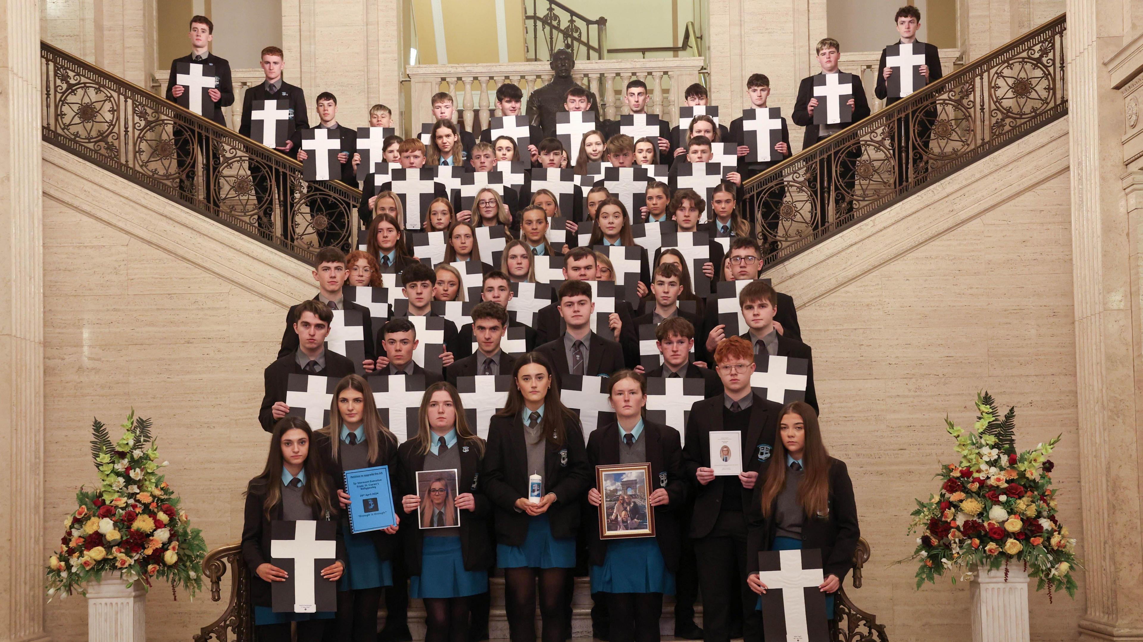 Pupils standing on steps in the Great Hall at Parliament Buildings. Each pupil, wearing dark coloured uniforms are holding crosses and photos of road crash victims