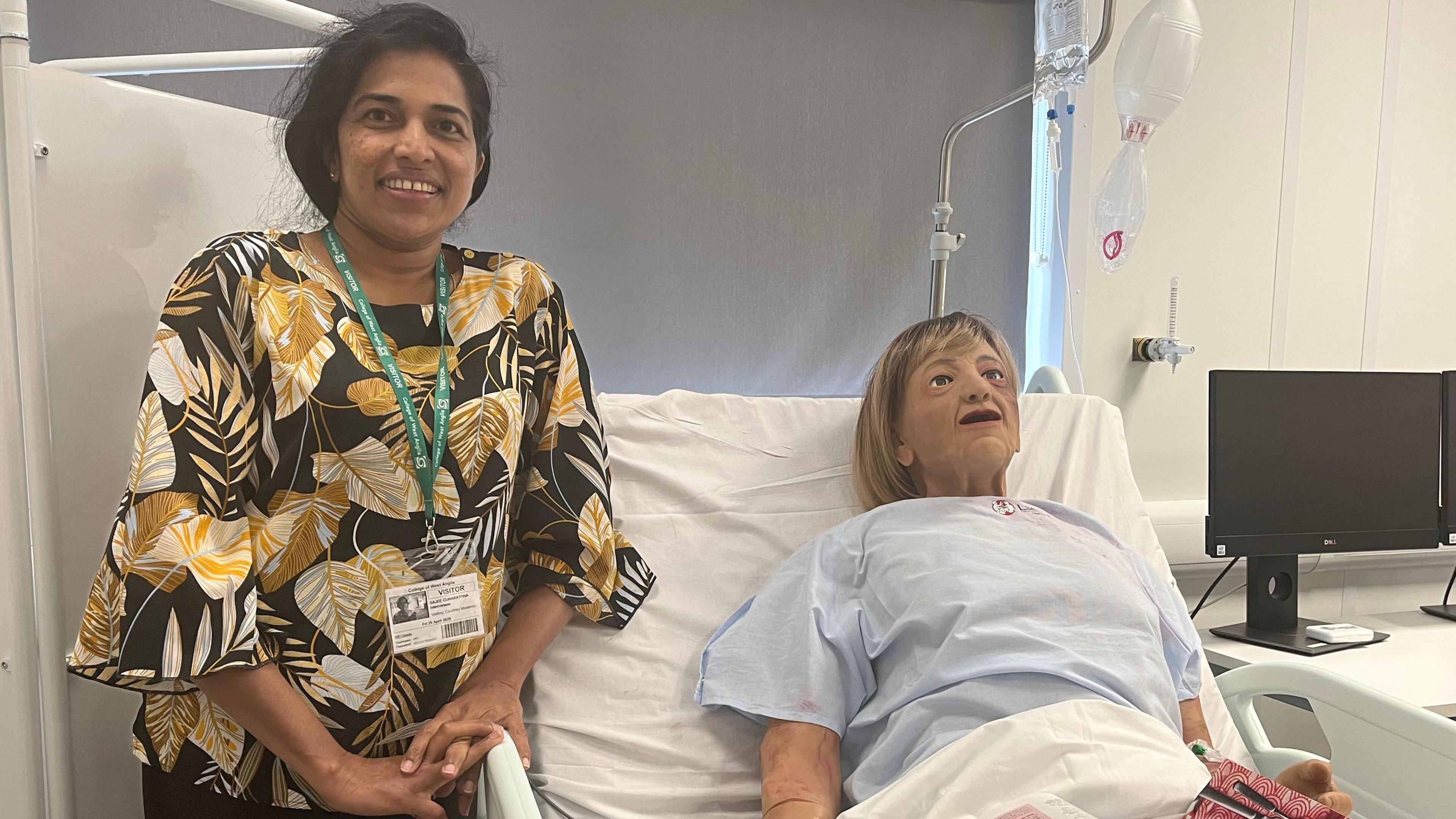 Senior care worker Sajee Gunarathna stands next to the bedside of a model patient called 'Dorris'. Sajee is wearing a black and yellow floral top and has long black hair.