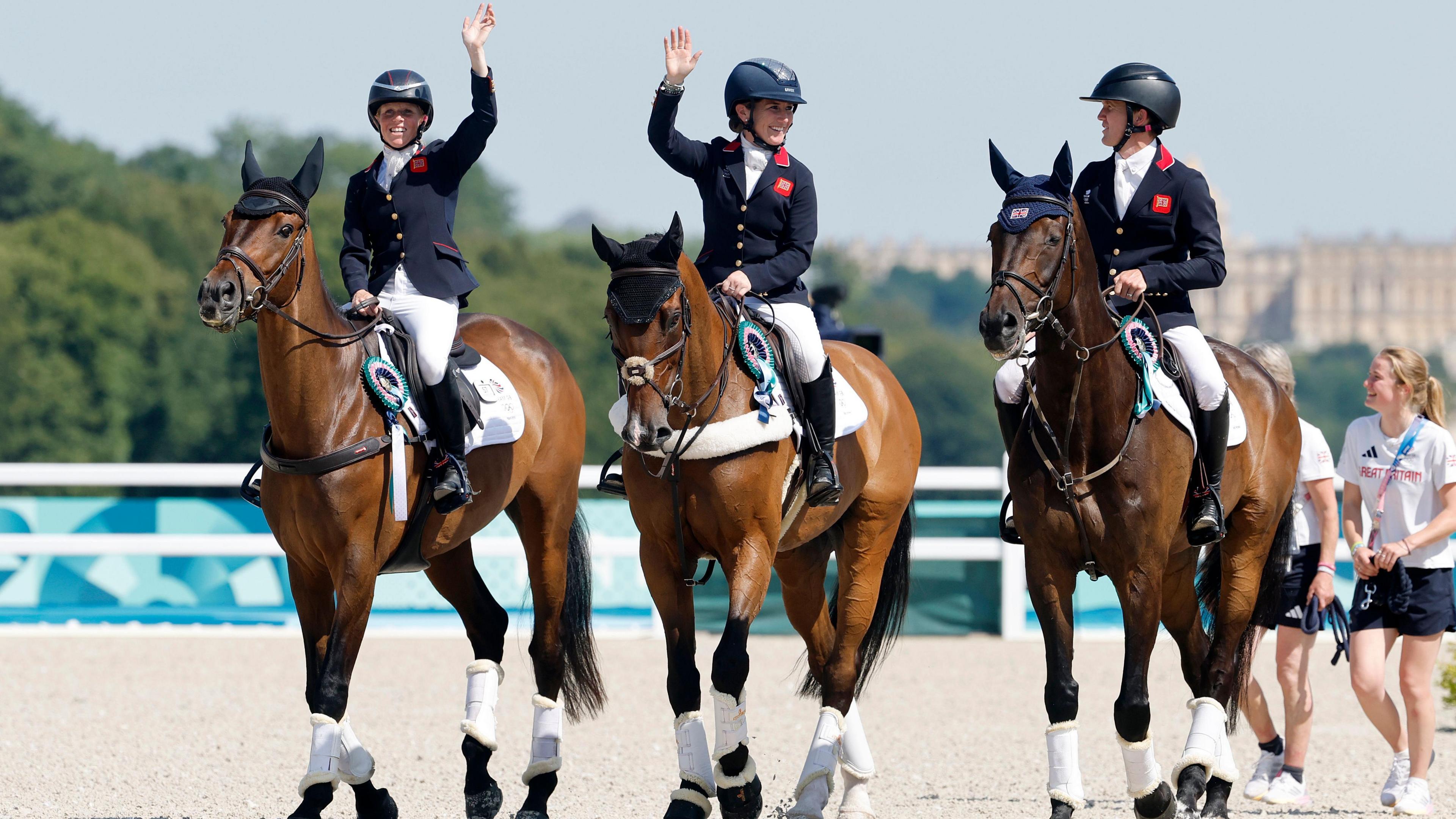 Ros Canter, Laura Collett and Tom McEwen wave to the crowds as they arrive at the medal ceremony on their horses, which are wearing rosettes.