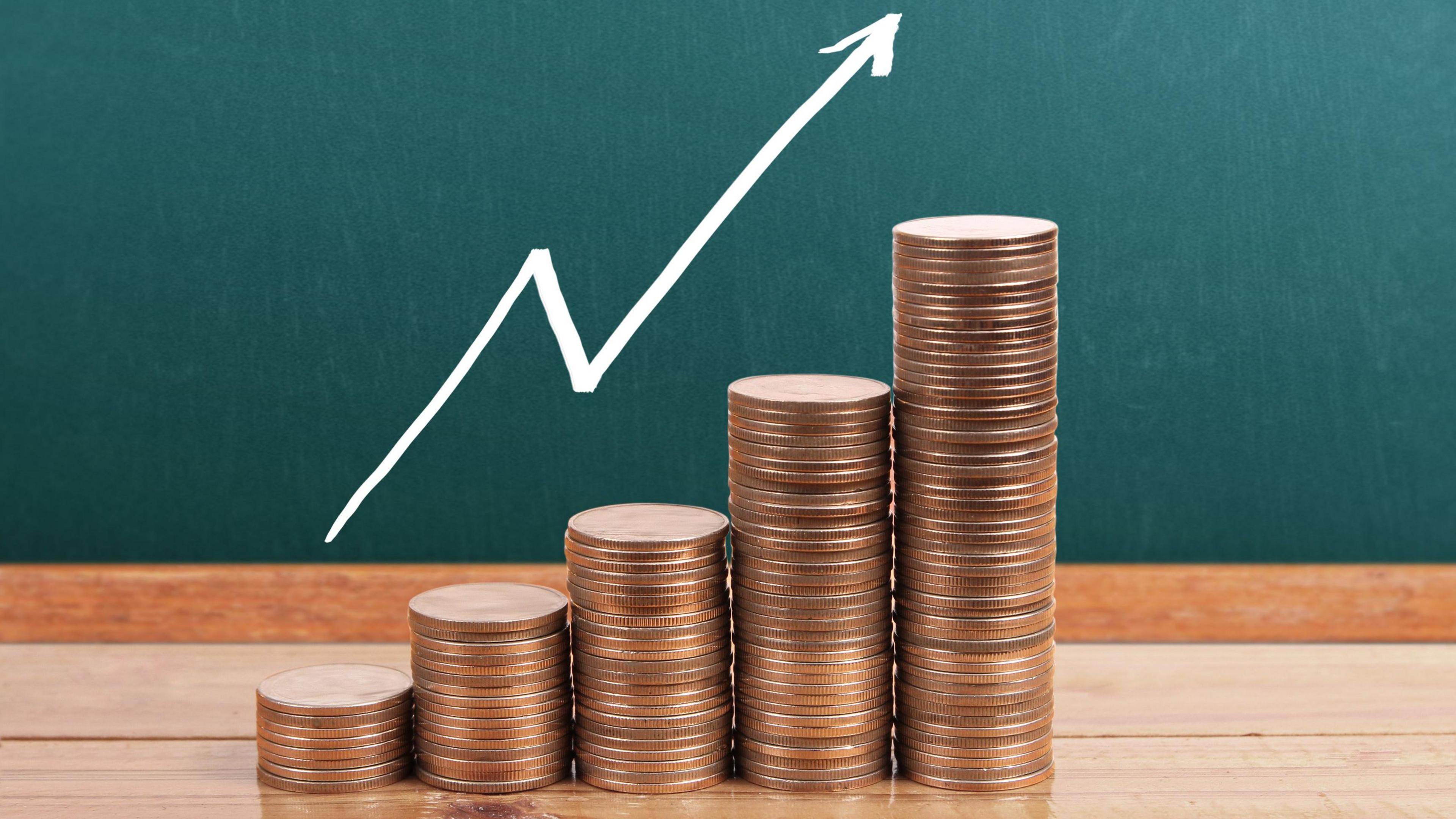 5 stacks of 5p coins that go up gradually with a white arrow signalling the amount of money going up. They are on a wooden floor and a teal wall is in the background.