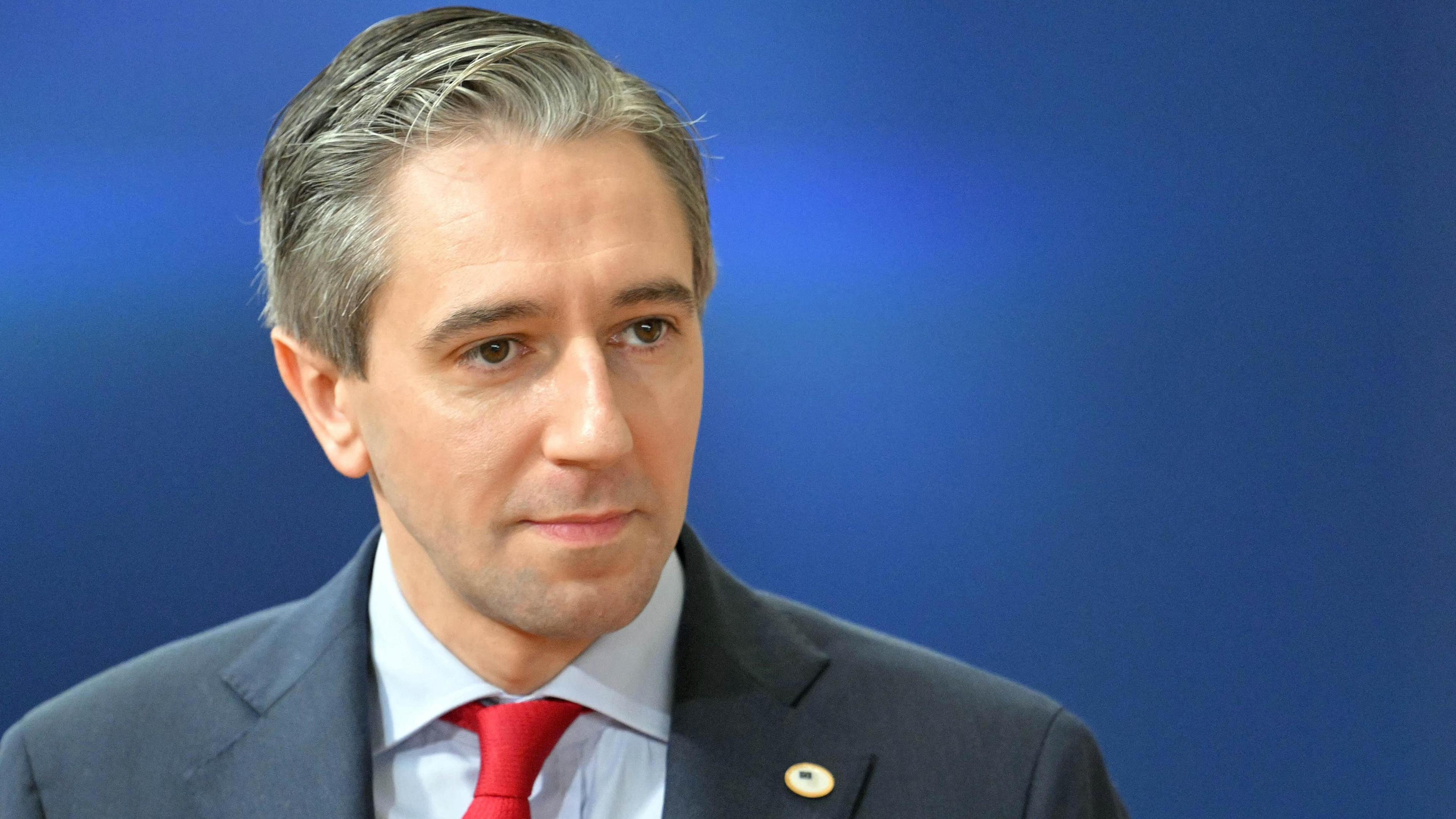 Simon Harris, who has short grey hair and wears a navy suit over a light blue shirt and a red tie, stands in front of a blue backdrop.