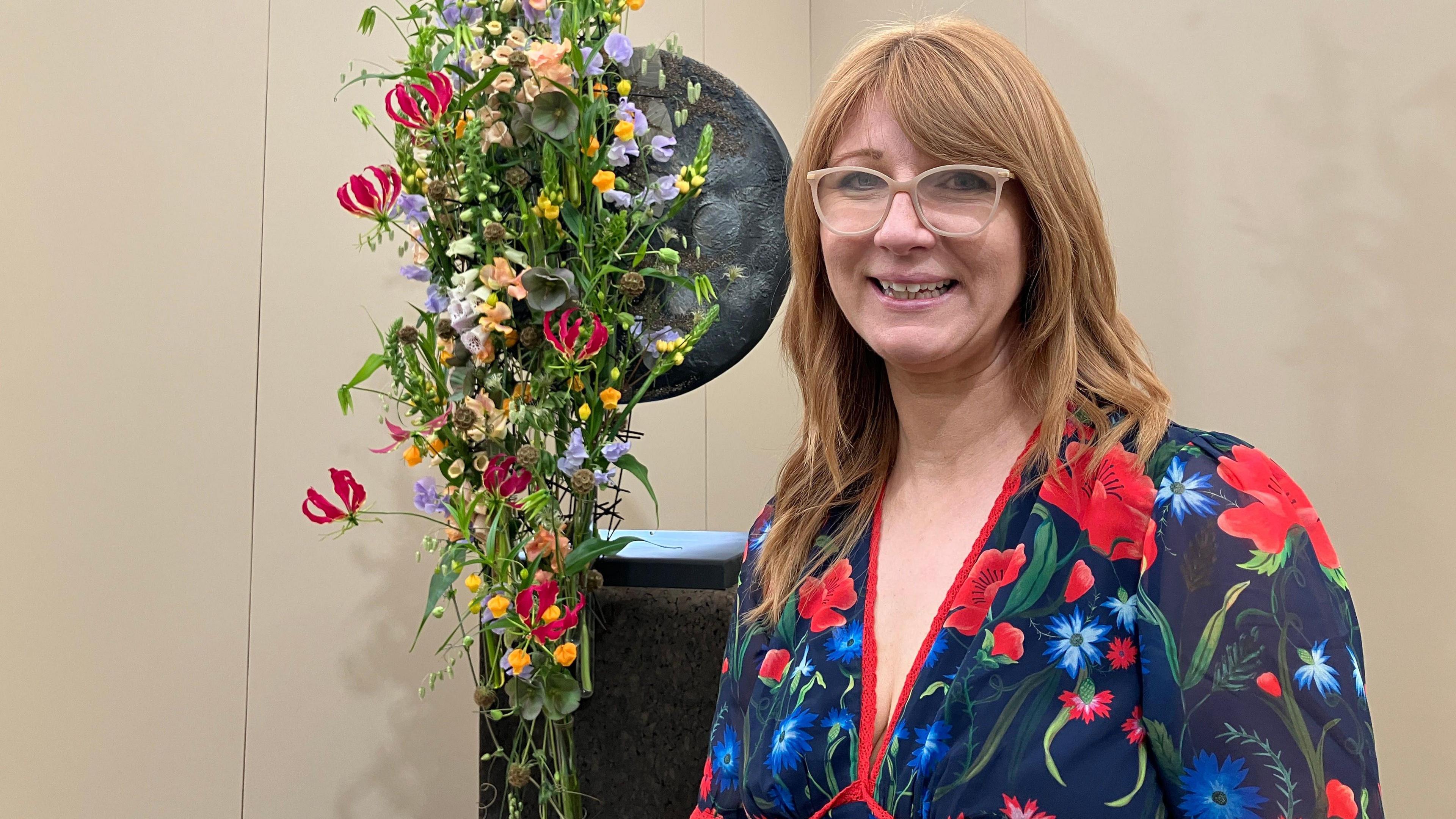 A bespectacled woman in a floral dress stands in front of a flower display.