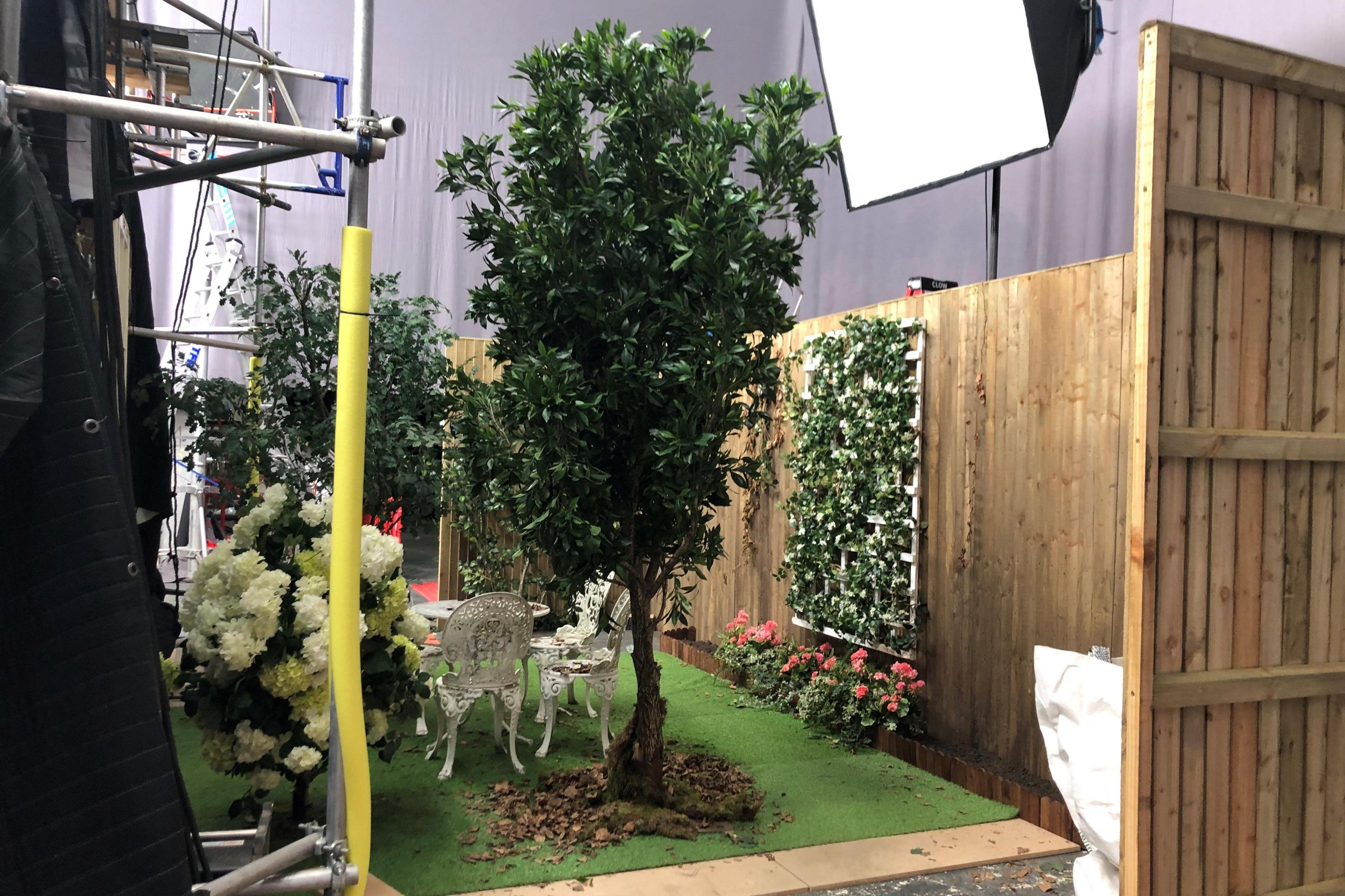 Plastic trees and flowers around a small fake green lawn with white chairs and tables in the middle and wooden fences around the sides