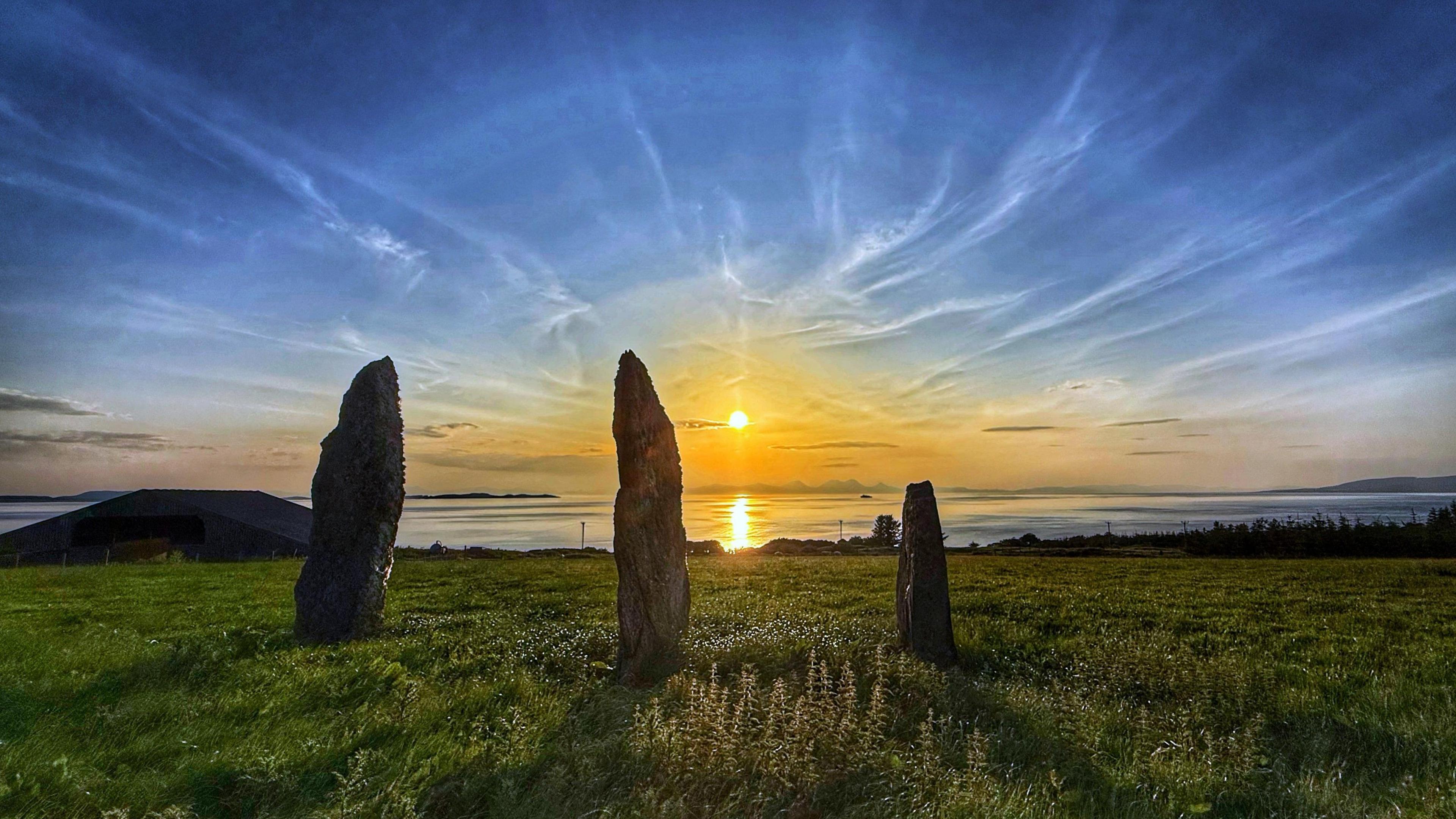 Three tall, slender stones standing upright in a grassy field at sunset. The setting sun casts a warm, golden light over the calm sea in the background, which is framed by a low-lying coastline. The sky above is a vibrant mix of blues and oranges, with streaks of cirrus clouds
