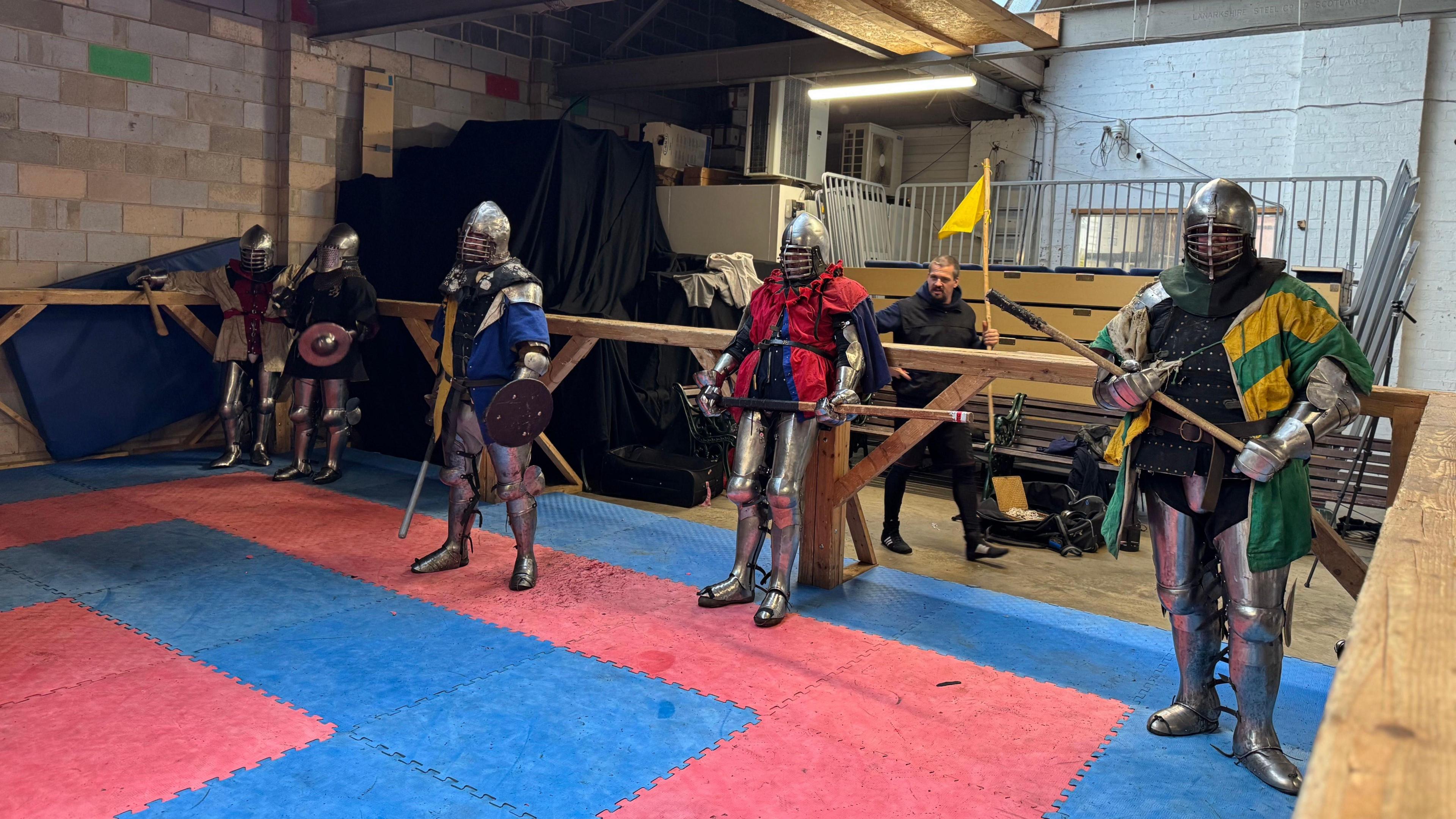 A group of men in armour lean against a wooden railing between rounds of their fight.