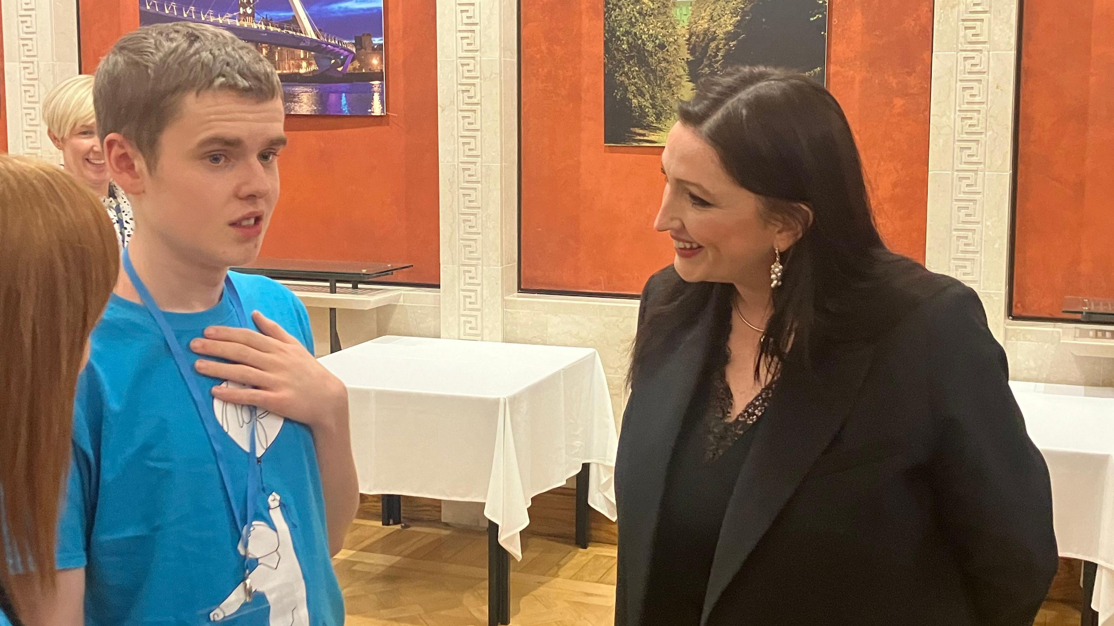 A photo of Deputy First Minister Emma Little-Pengelly smiling as she talks to Caleb White on her right. Ms Little-Pengelly is wearing a black suit jacket and black top. Caleb is wearing a blue shirt with a white logo of a boy holding a balloon.