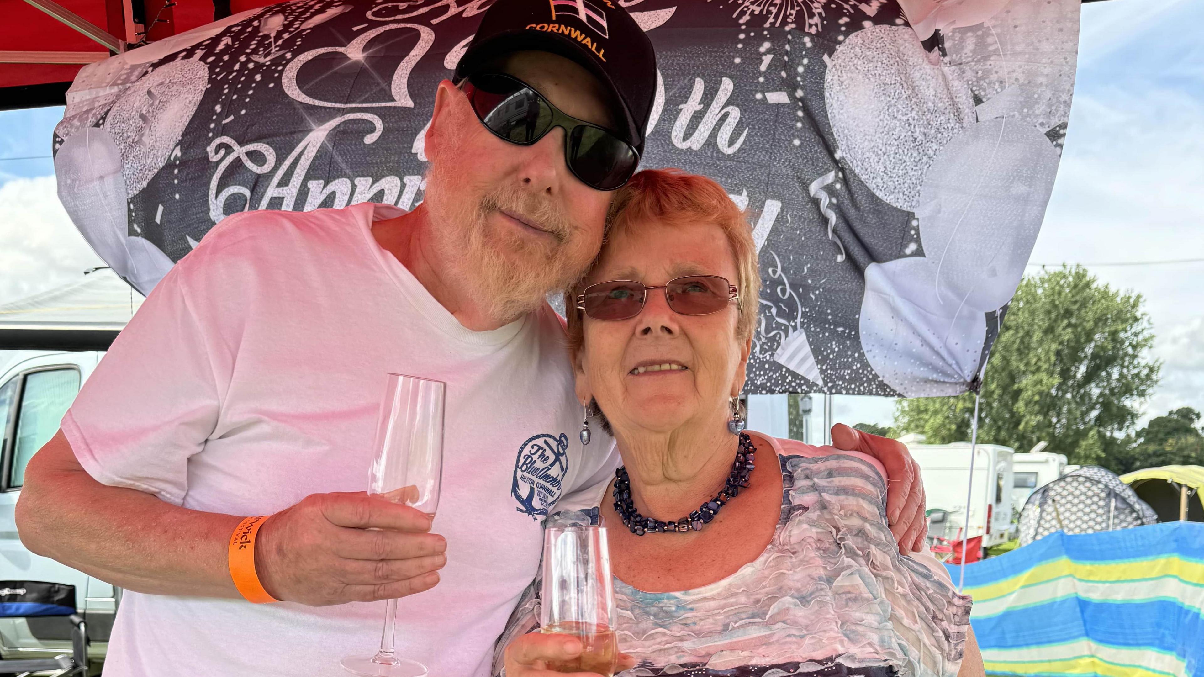 Mary and Derek Droscher standing together behind a banner to celebrate their anniversary. Mary is wearing sunglasses and holding a glass of wine. Derek is wearing dark sunglasses, a black cap and holding a glass of wine