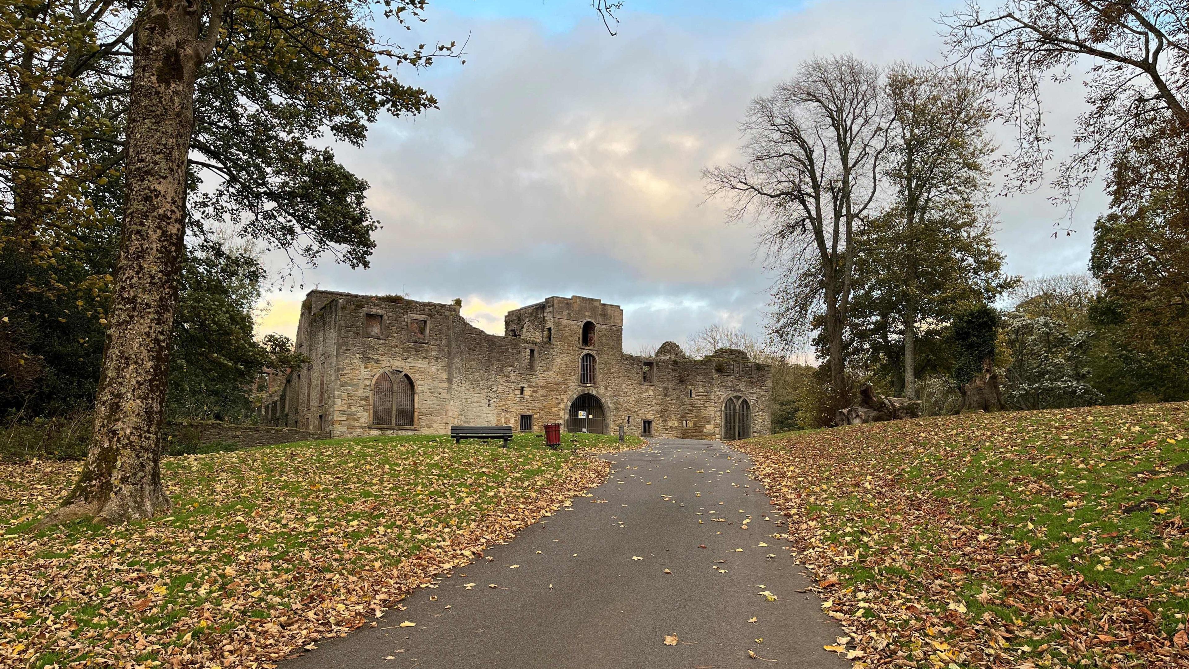 Workington Hall. The ruin of the castle has no roof and bars at its widows. A tarmacked footpath leads up to it. It's surrounded by grass covered in fallen leaves from the nearby trees.