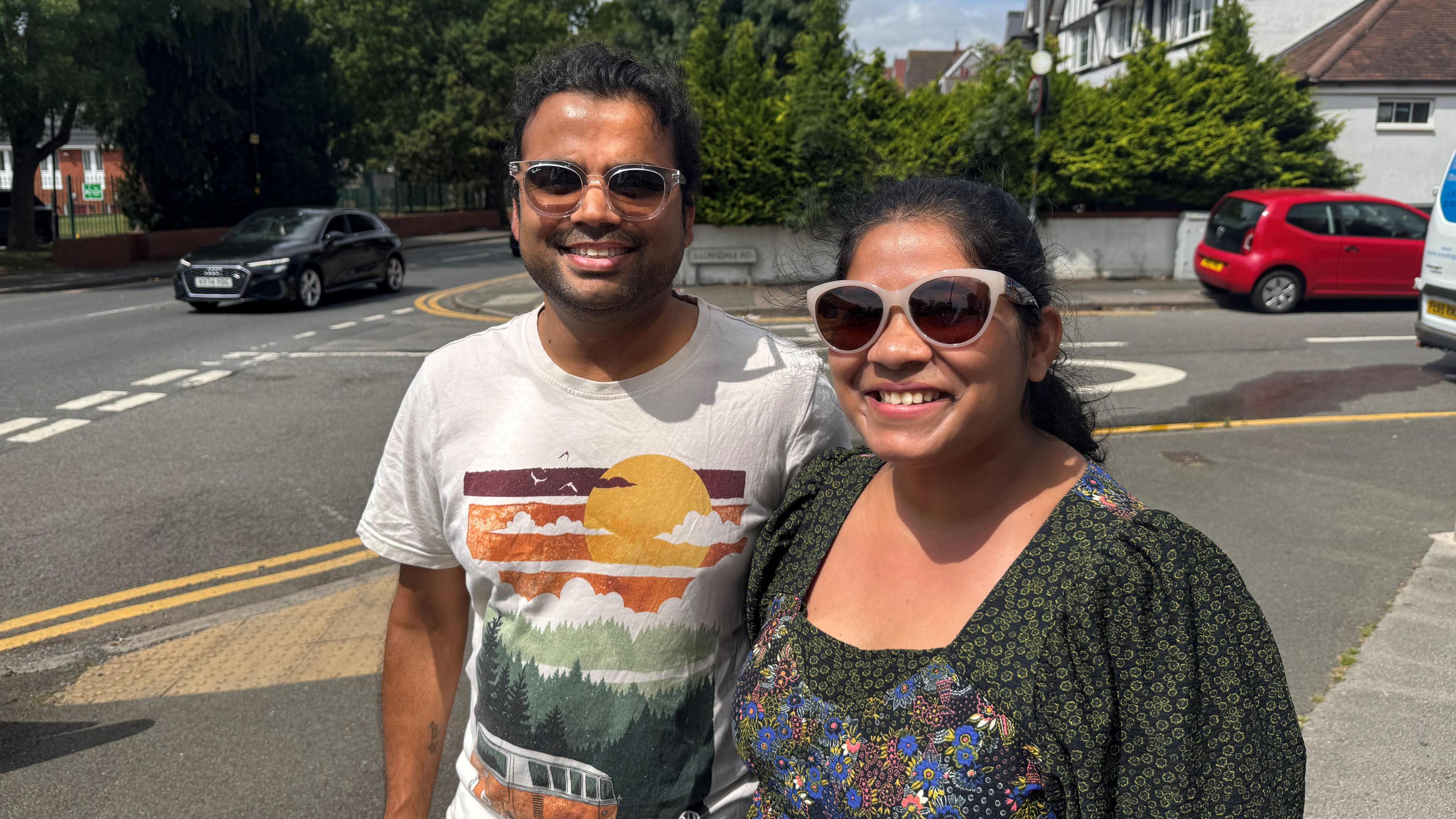 Husband and wife Amar and Sonal Mundada. They are standing at the junction of a busy road. They are both smiling and wearing sunglasses. Amar has an arty T-shit on. Sonal is wearing a green blouse.