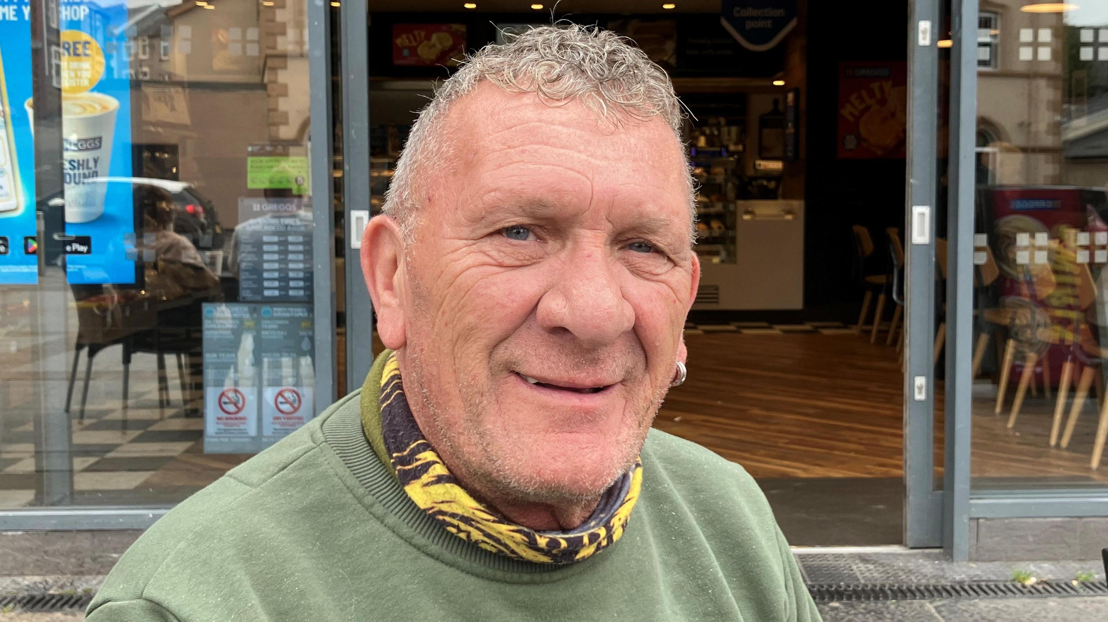 A man with salt and pepper, short curly hair wearing a green sweatshirt and a yellow and black snood. There are the doors to a cafe behind him. It is a head and shoulders shot.