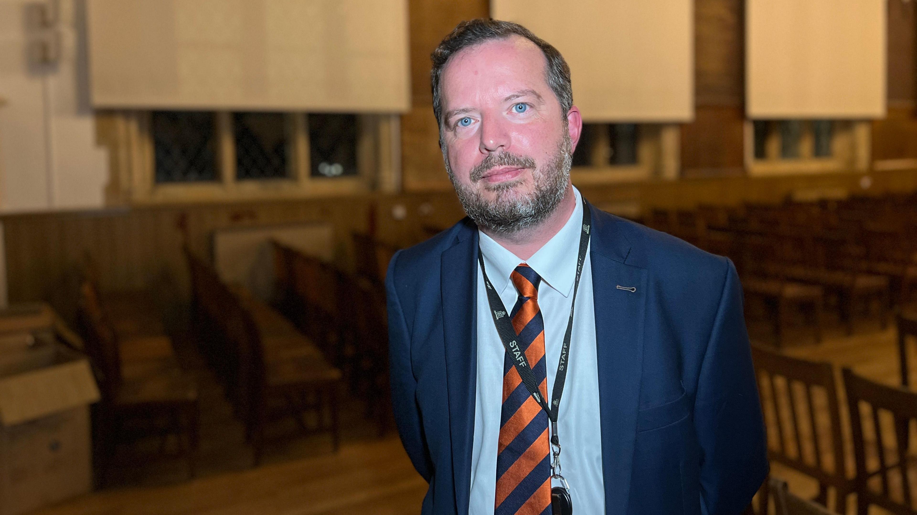 Anthony looks at the camera while standing next to chairs in a empty school hall. He is wearing a navy blue jacket with a light blue shirt underneath and a orange and blue stripped tie. He has short grey hair which is brushed to the side.