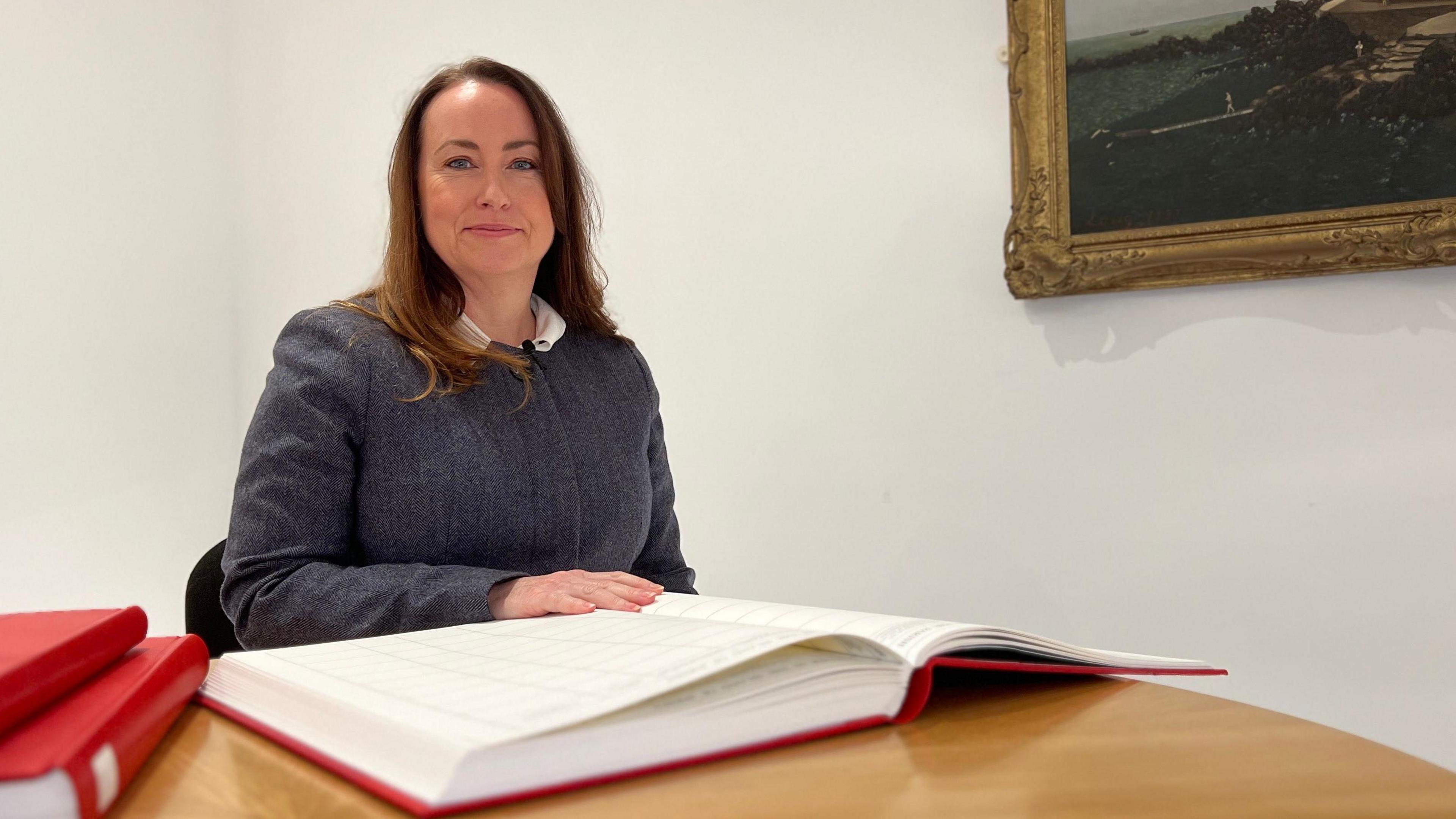 Claire is seated at a wooden table indoors with an open large ledger book in front of them and two additional red-covered books stacked nearby. A framed painting of a coastal scene hangs on the white wall behind them.