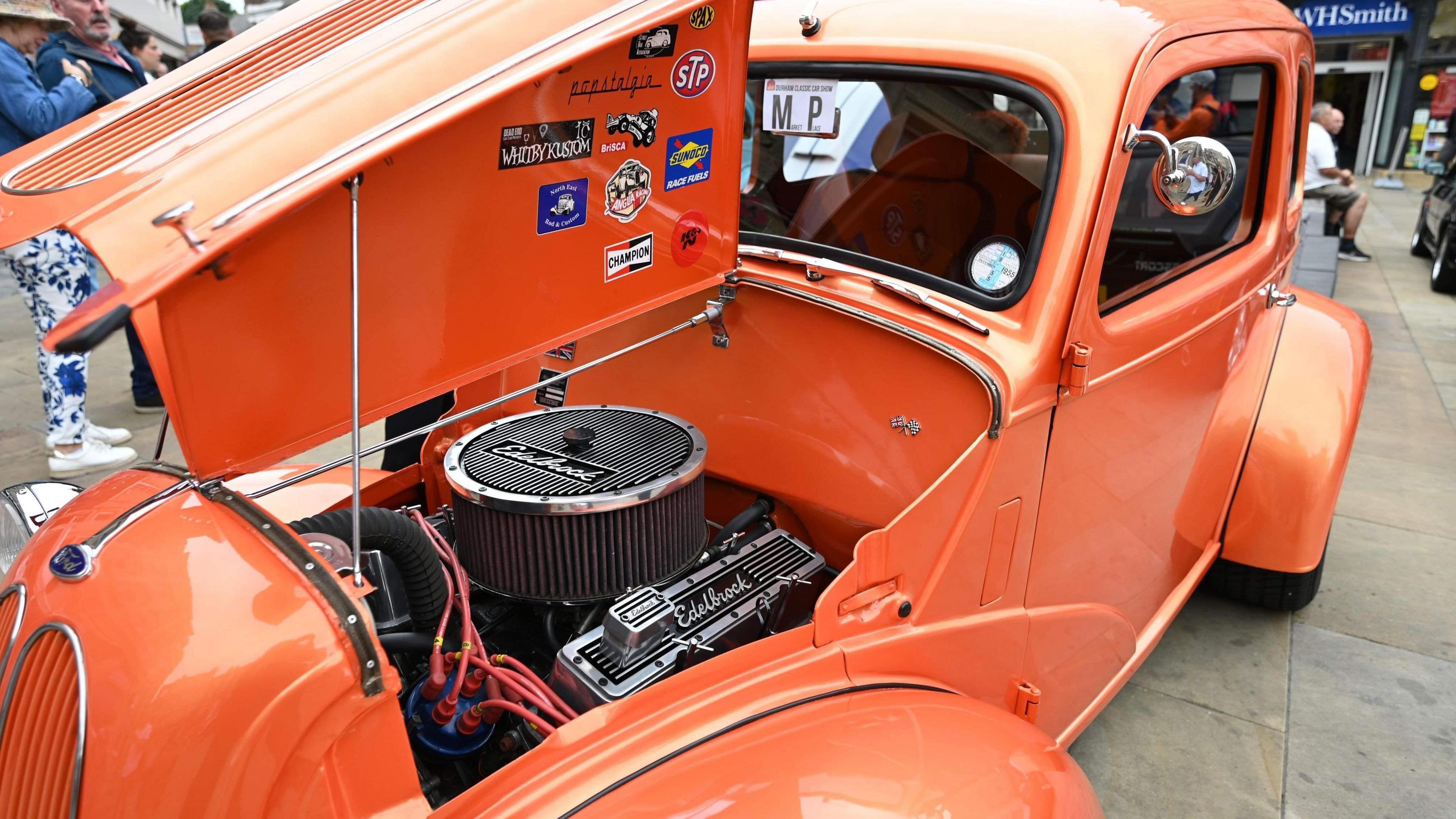 Close up on the front of an orange vintage car with its bonnet open showing its engine