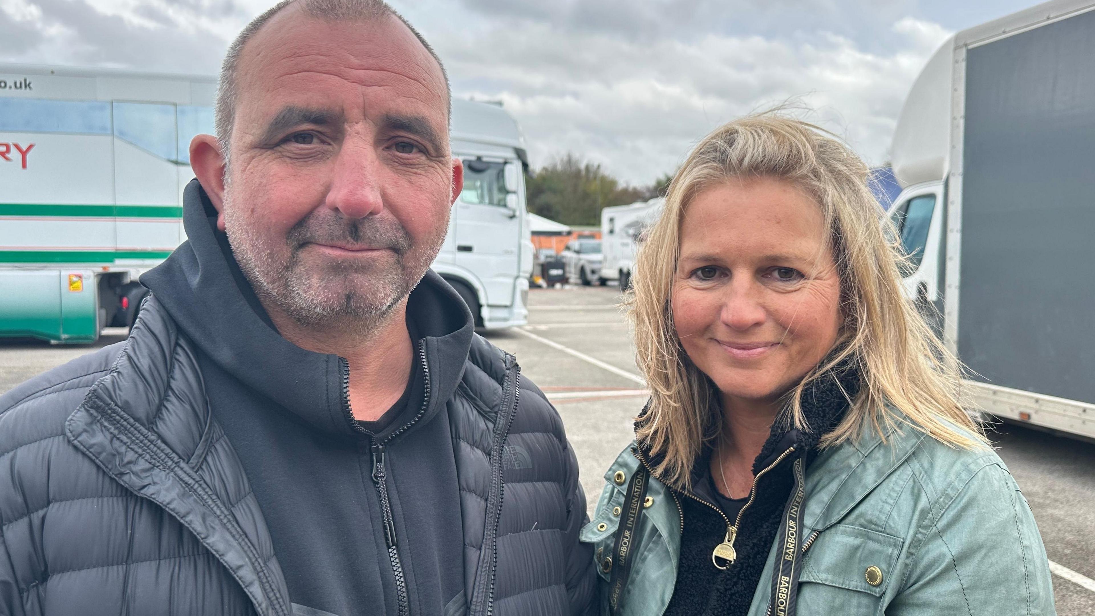 Man with short hair and stubble with a dark hooded top and jacket standing next to a woman with long blonde hair, wearing a dark fleece under a mint coloured jacket. There are two large vehicles in the background.