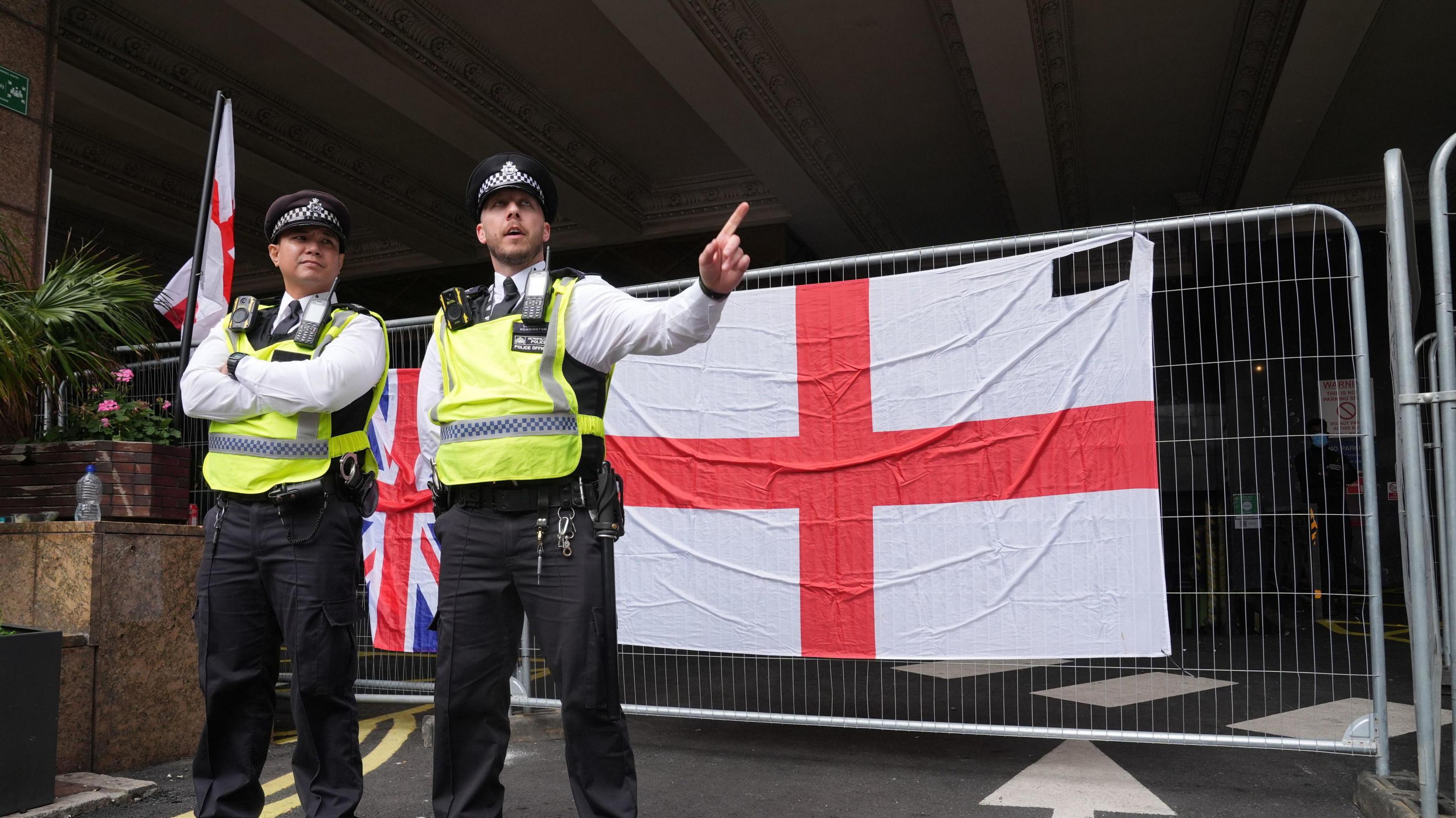Two police officers in high-vis jackets standing in front of a temporary security fence with an England flag draped over it in Canary Wharf, London.