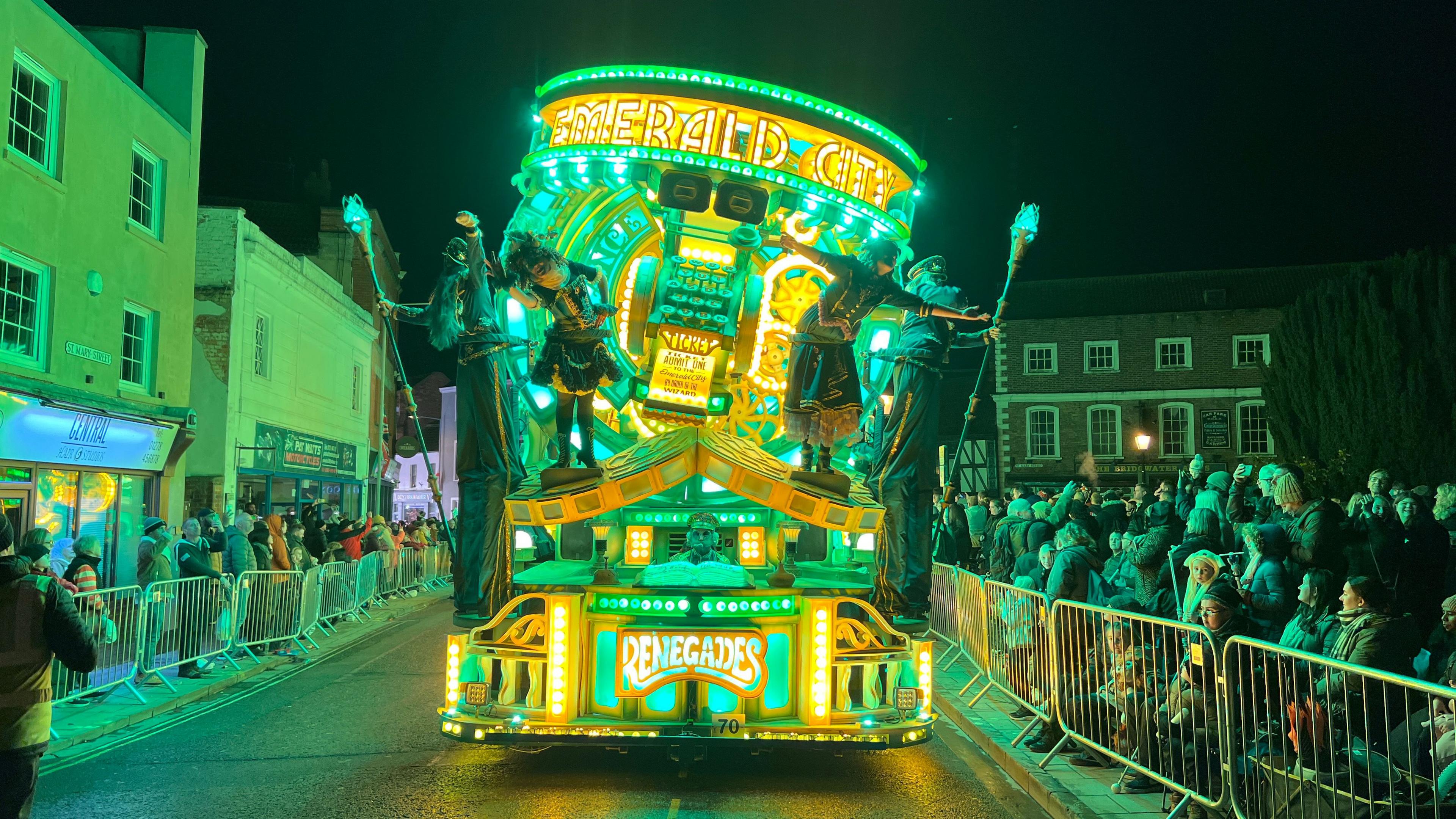 A Renegades Carnival Club float is being driven down a street in Bridgwater. The float is illuminated with bright yellow and green lights. The words EMERALD CITY are written on the top of the float, and the RENEGADES logo is on the bottom front. There are four carnival performers at the front of the float. Dozens of spectators are behind barriers watching the float go by.