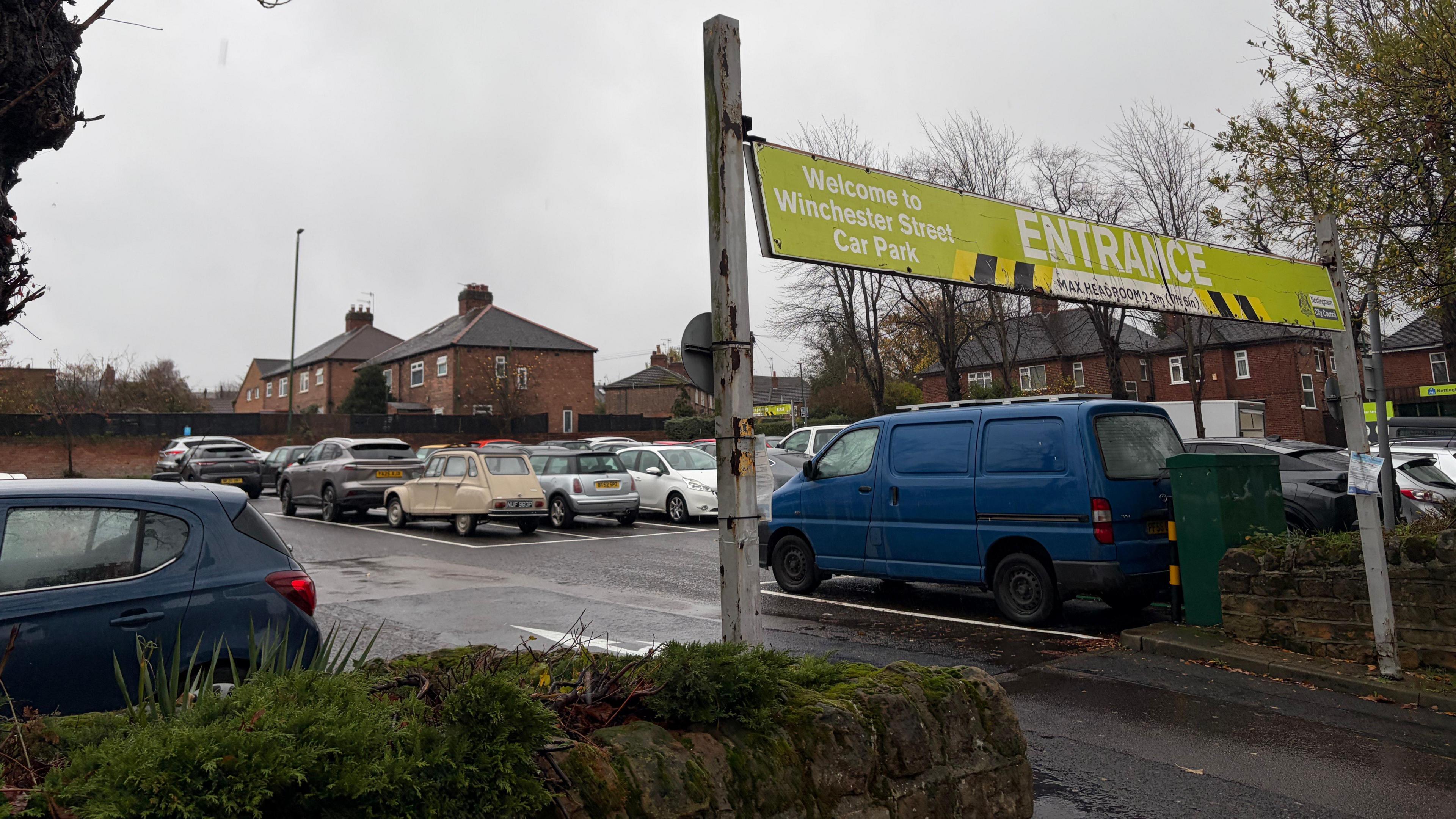 An entrance sign leading to a car park with rows of cars lined up