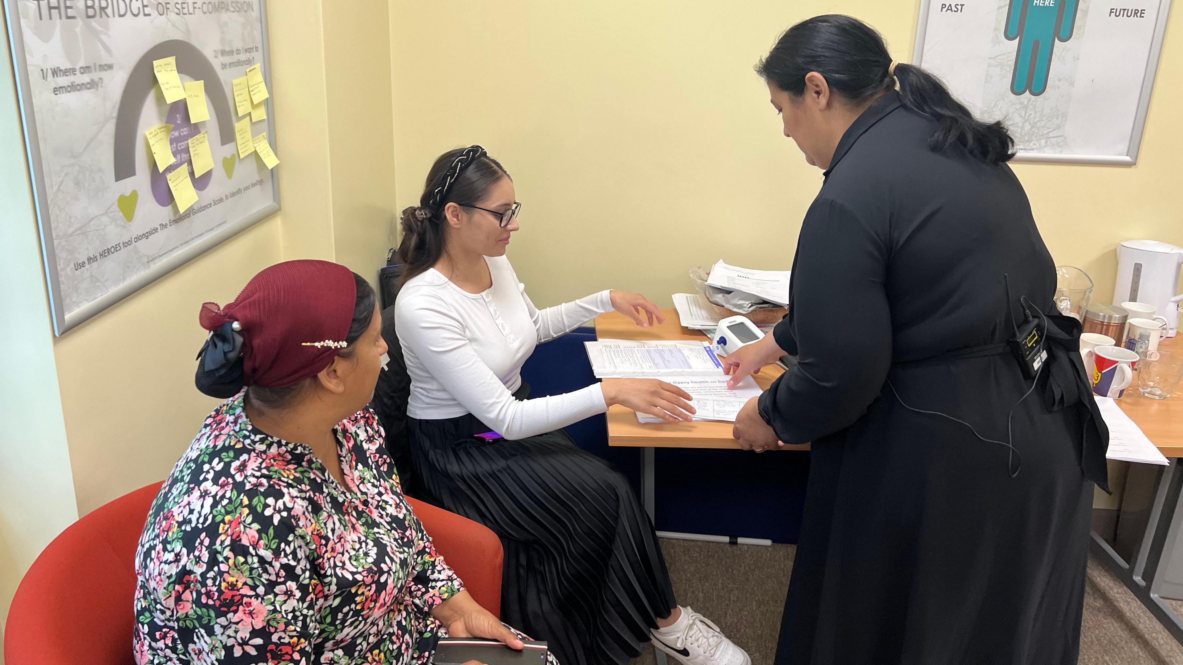 In a room with a table and posters on the wall, two women are seated and a third woman, wearing a black dress, is standing and showing one of the women some paperwork on a table.