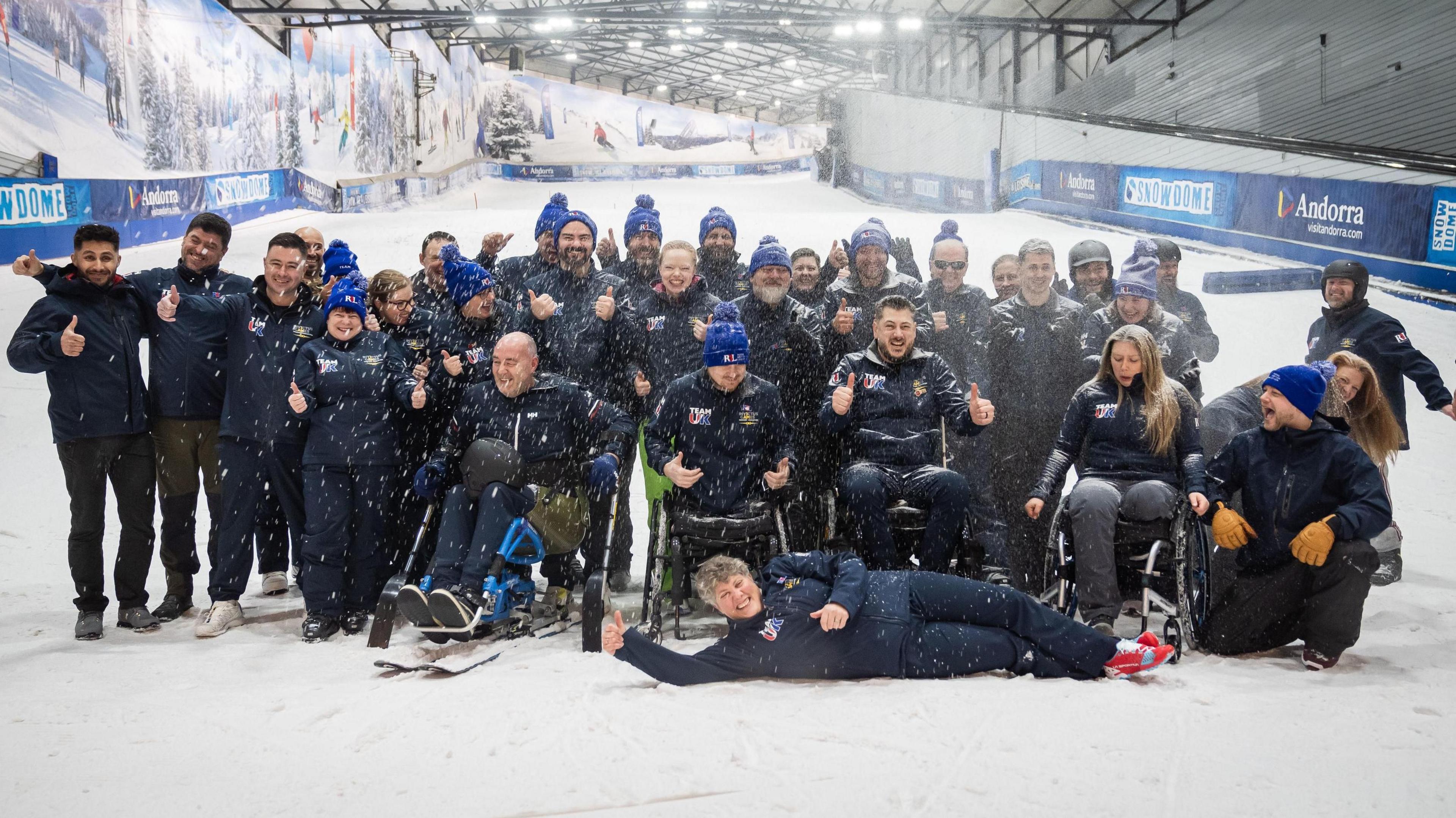 A group of people at the bottom of an indoor snow slope. They are all wearing branded team tops and four of them are in wheelchairs.