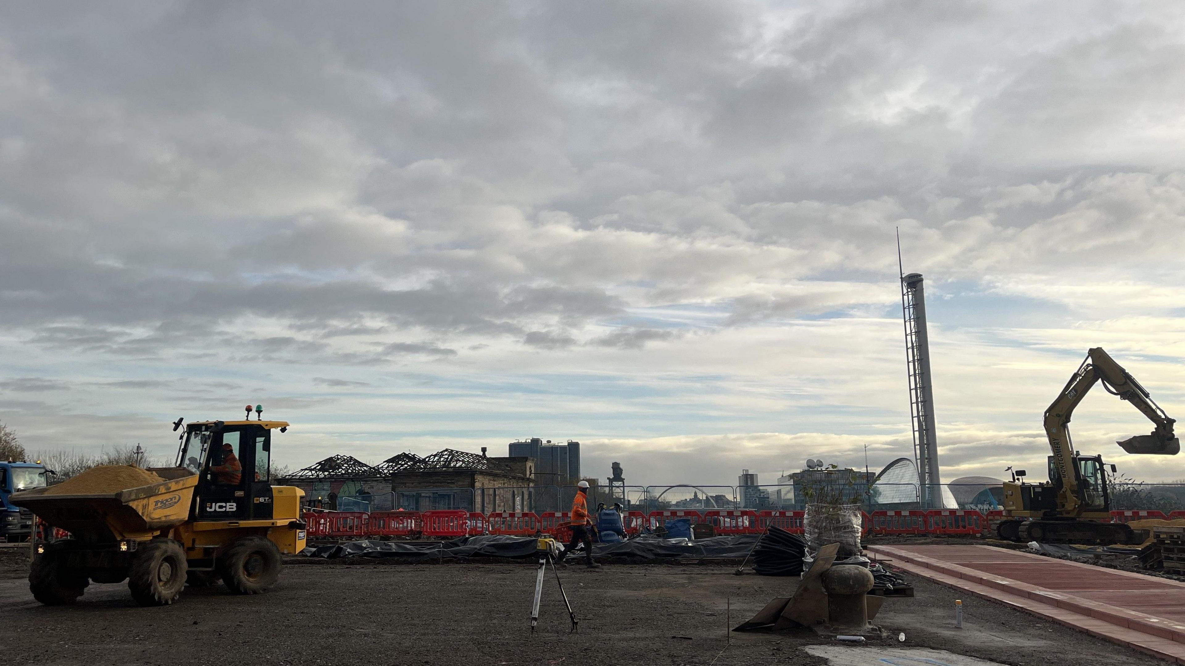 The building site for the new park. There are two diggers and workmen on the site. The pump house, the Science Centre and bridges over the Clyde can be seen in the background. 