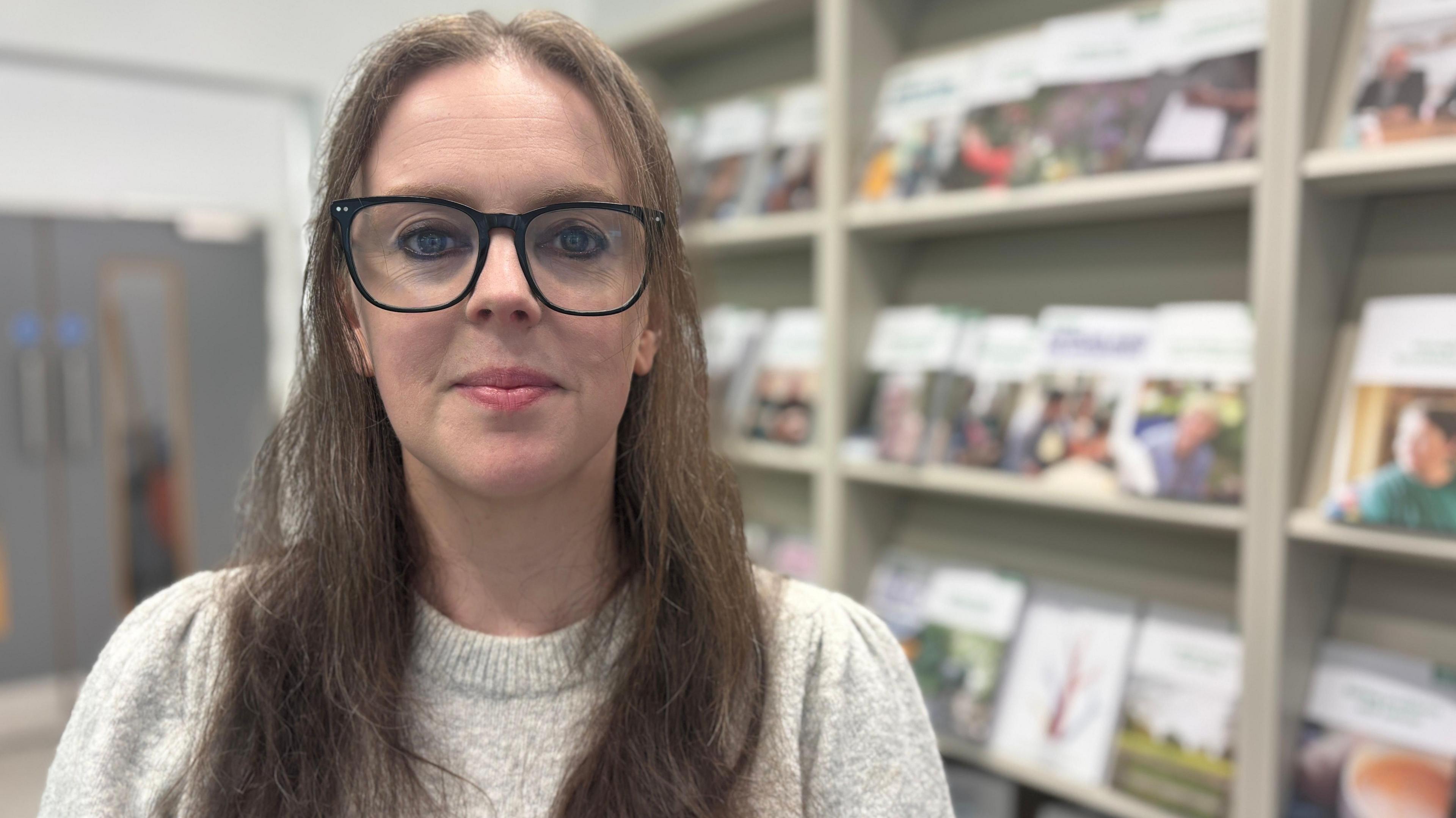A young woman stands in front of a wall of books and leaflets. She is wearing glasses and a pale grey jumper. Her long brown hair is partially pinned back.