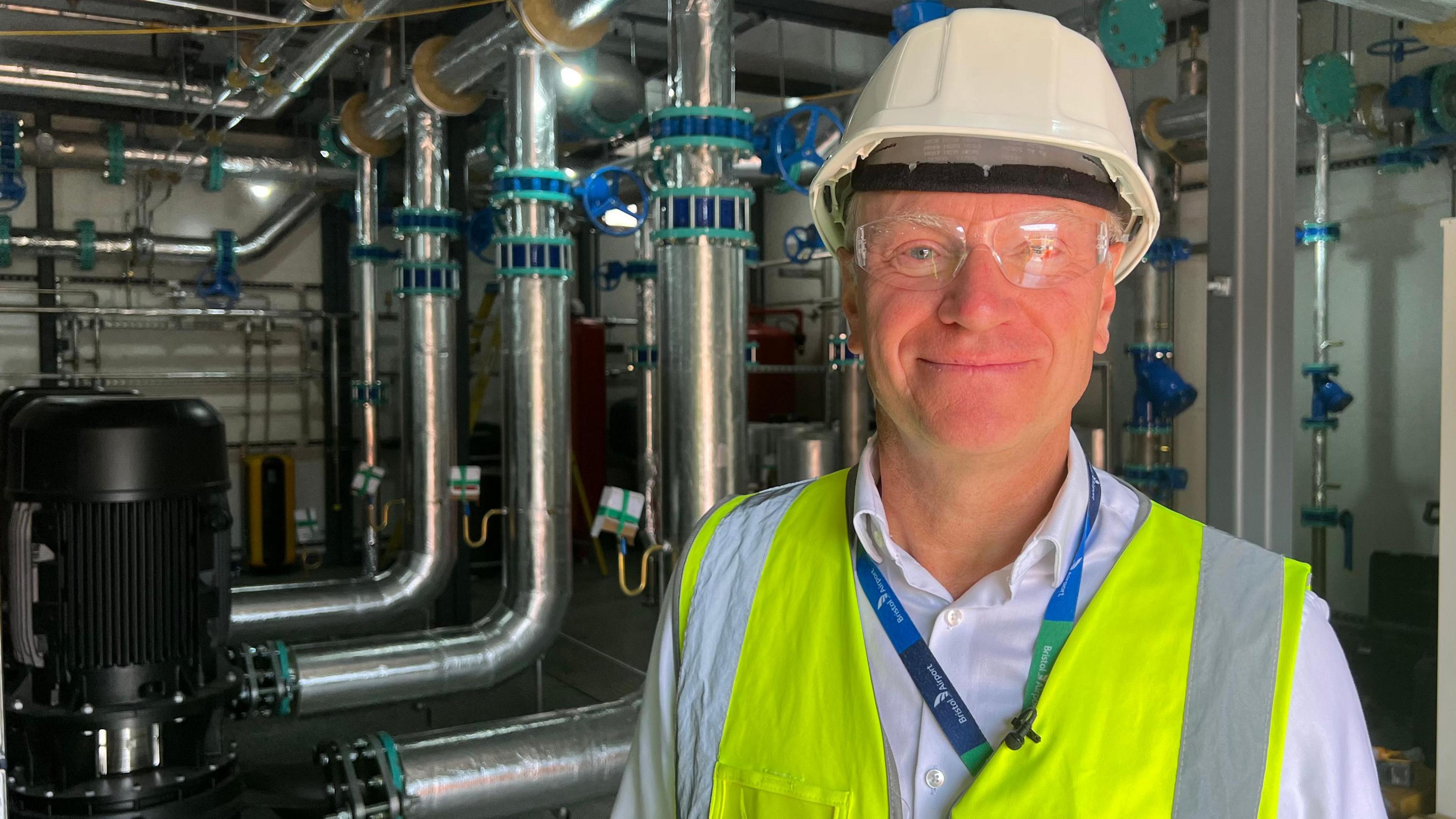 Dave Lees is wearing a hard hat and high-vis jacket and goggles while stood in a room with silver pipes behind him.