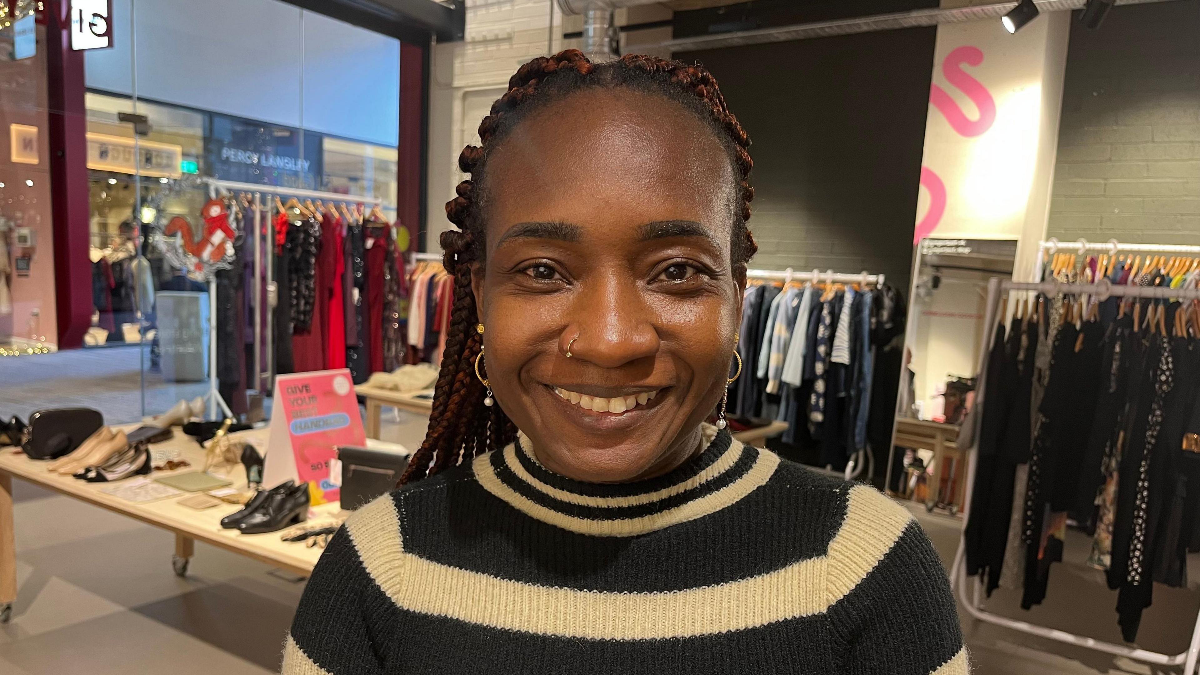 Kemi Ogunlana, a woman with long red and black hair worn in braids and wearing a black and white striped knitted dress. She stands in front of tables and racks of clothes