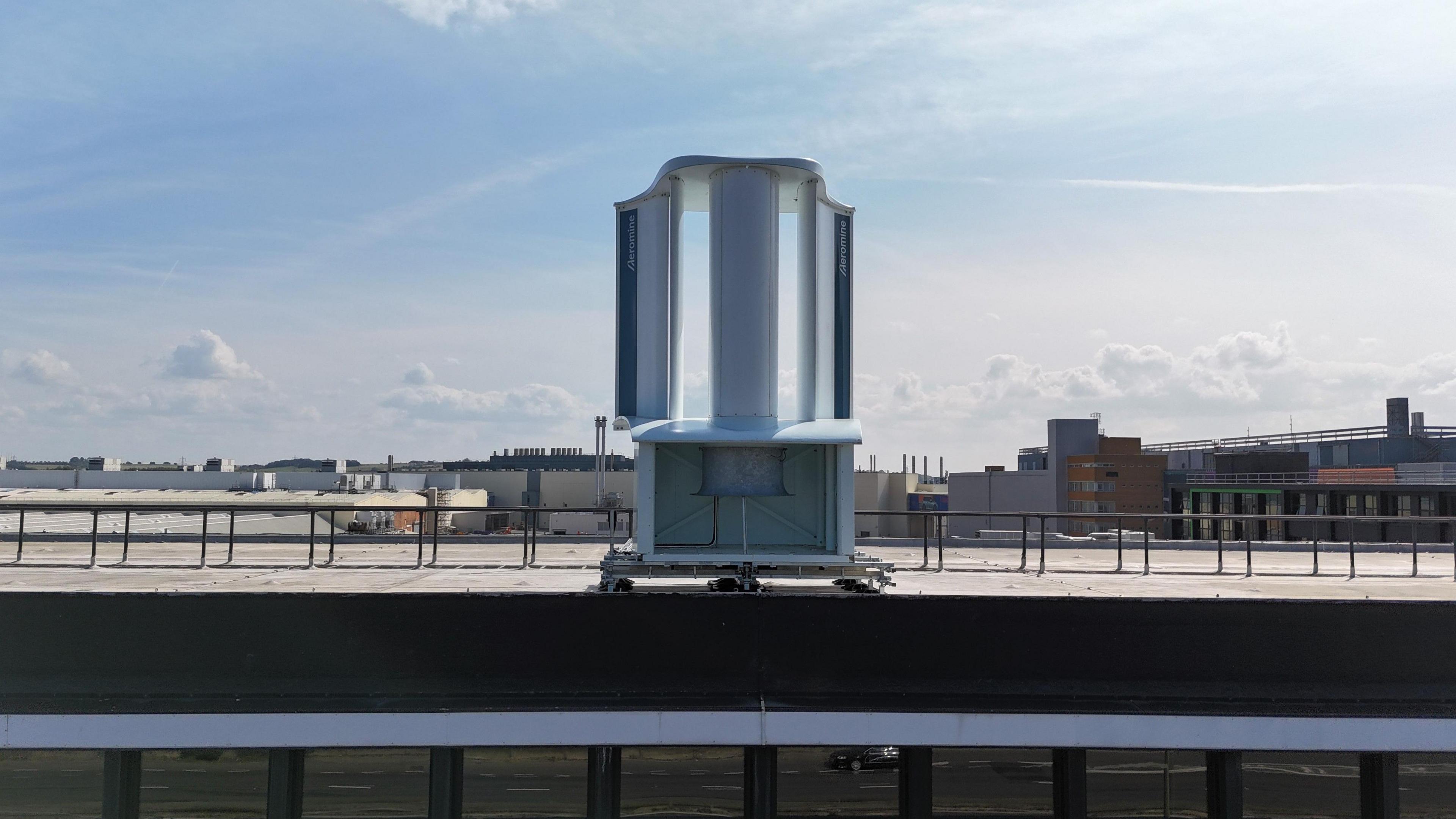 The new wind energy unit on top of the Oxford's Mini plant. It sits on the edge of the building. Other building can be seen in the distance. It's a cloudy day.