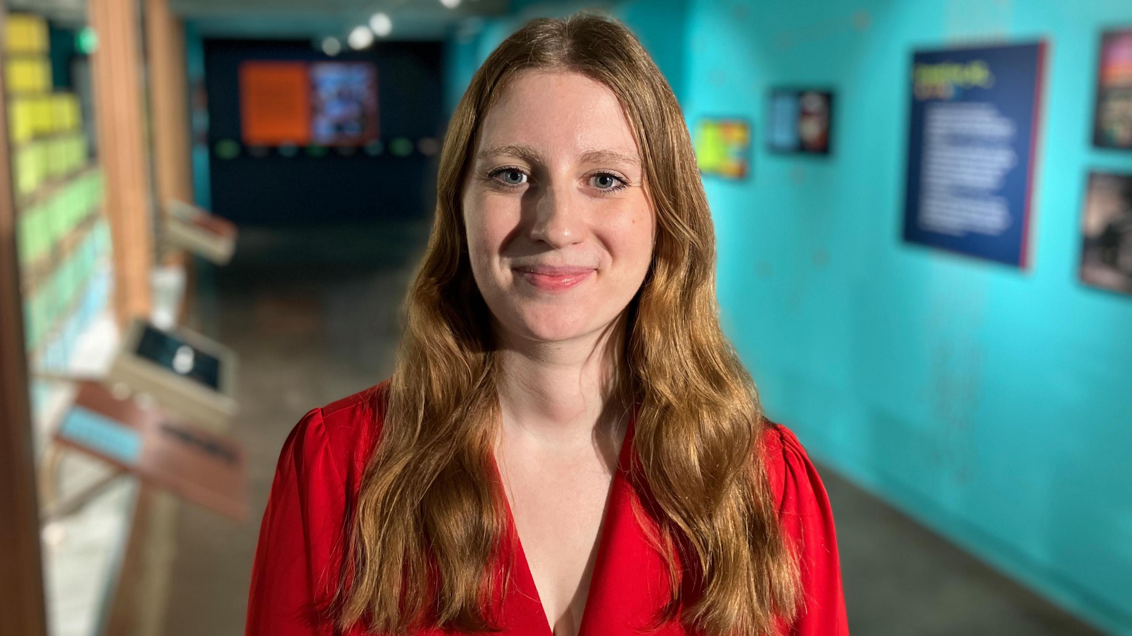 Tilly Davis has long auburn hair and is wearing a red blazer. She is stood smiling close to the camera at the exhibition