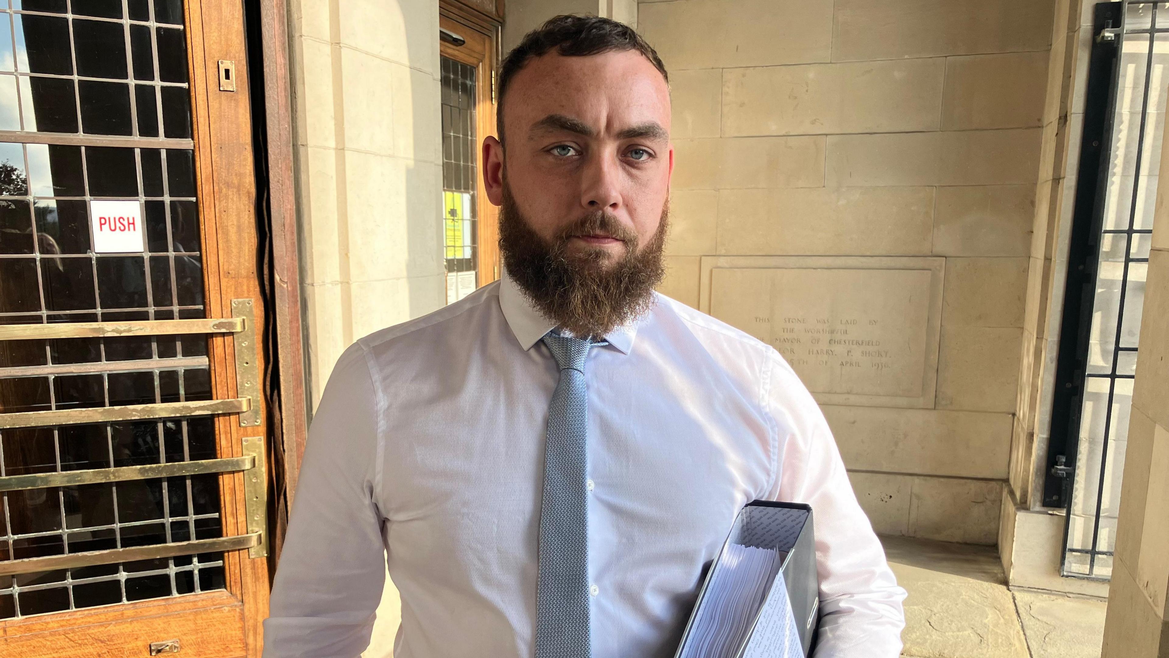 Jerome Allford wearing a white shirt and sky blue tie holding a folder looking into the camera. He has short hair and has a light brown beard.