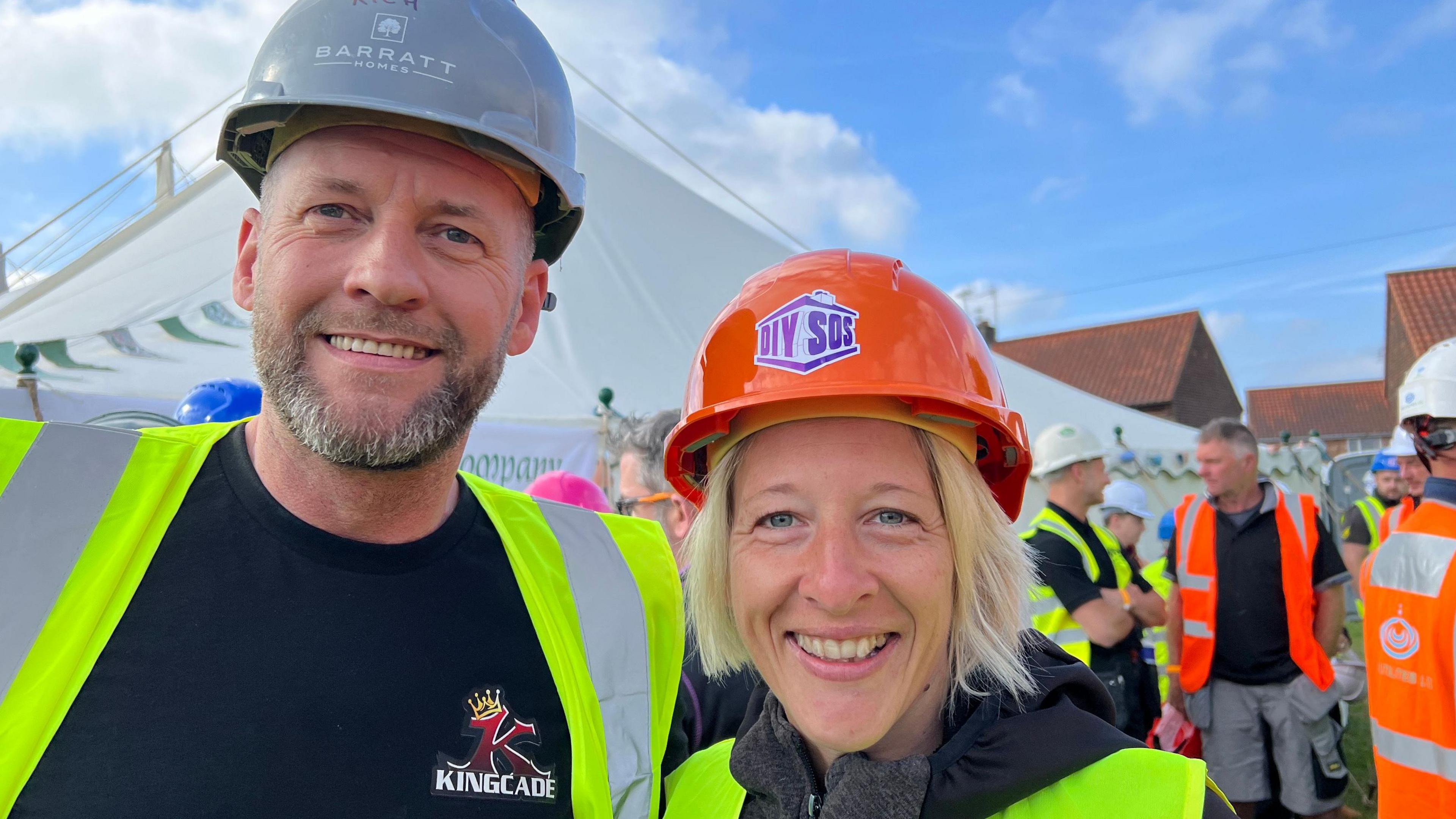 A man and a woman smile into the camera while other tradespeople gather in front of a marquee tent in the background. The man is wearing a grey hat, yellow safety vest and a black t-shirt. The woman, on the right of the image, has blonde hair and wearing an orange helment with a DIY SOS logo on the front.