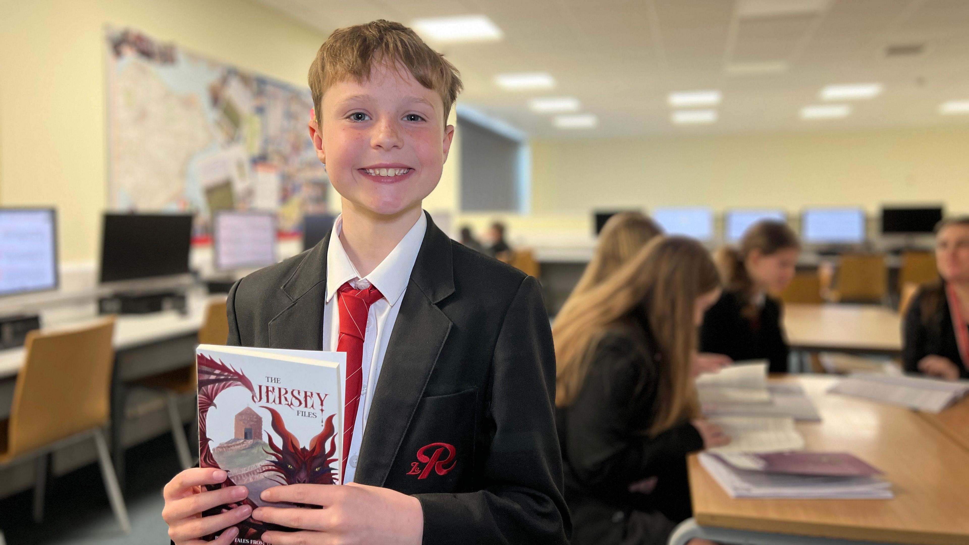 Ned smiles at the camera as he holds the book in his hands. He is wearing a black jacket with the school logo on his chest. Ned has short brown hair and a red tie. Behind him other pupils are reading the book on a table in a library.