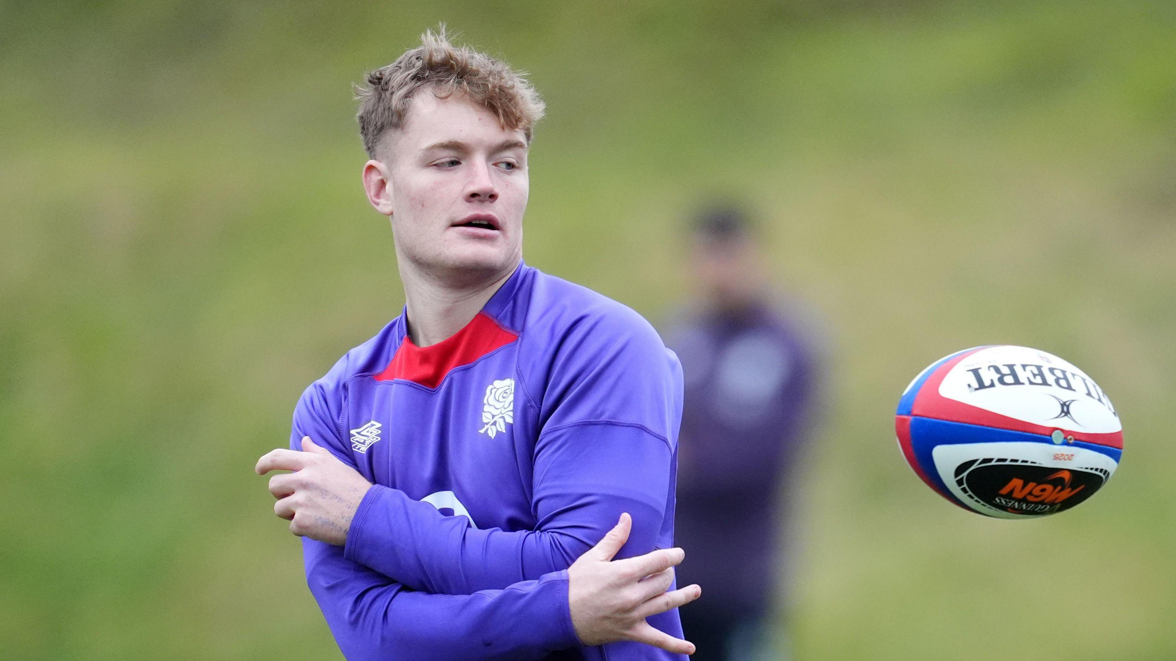 Fin Smith is in a blue training top and is throwing a rugby ball to the right. 