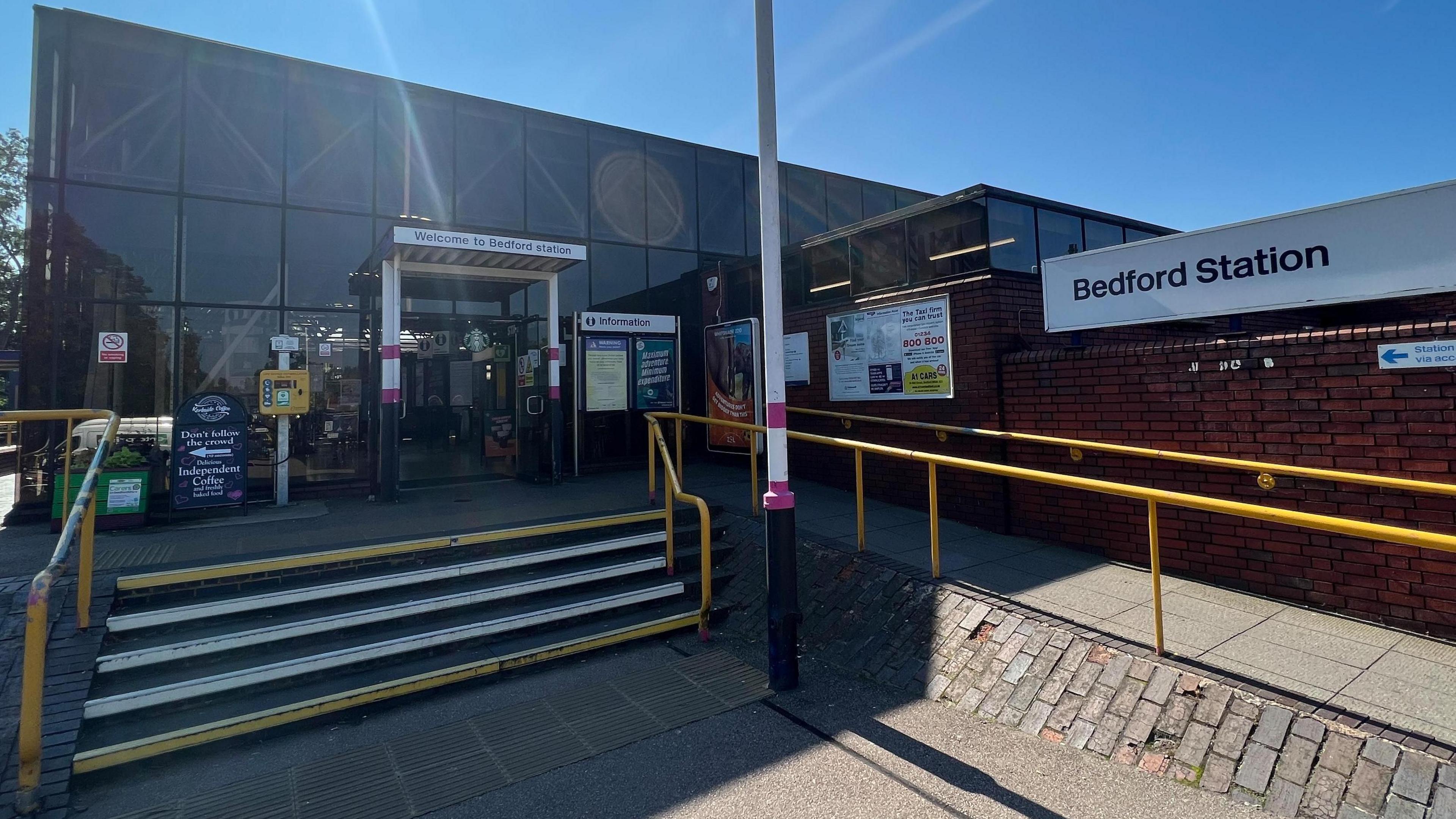 The front of Bedford Station with steps and an accessibility ramp leading to the glass fronted entrance.