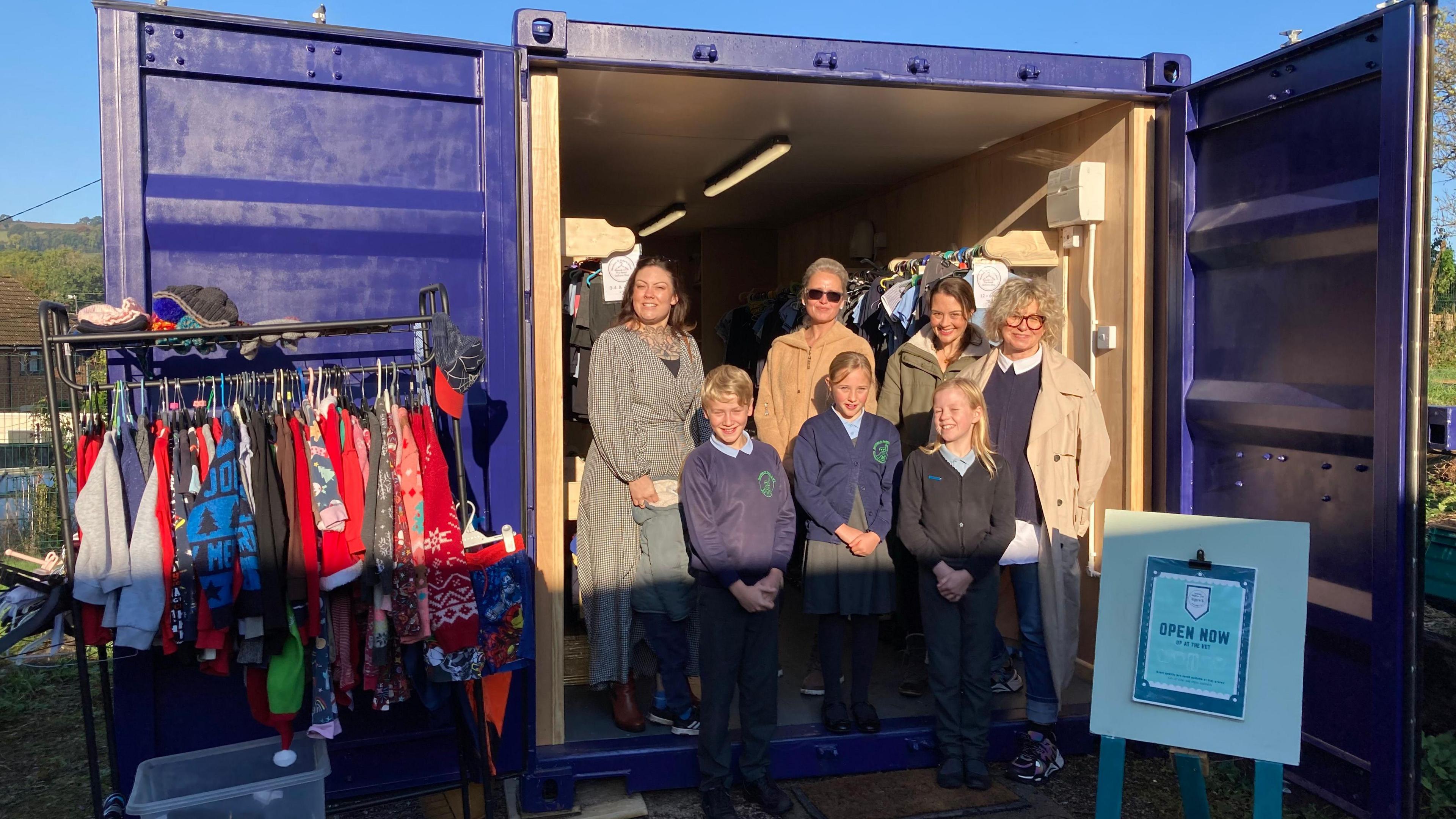 Parents and children outside a shipping container with second hand uniforms inside and a clothes rack just outside on the left hand side of the door.